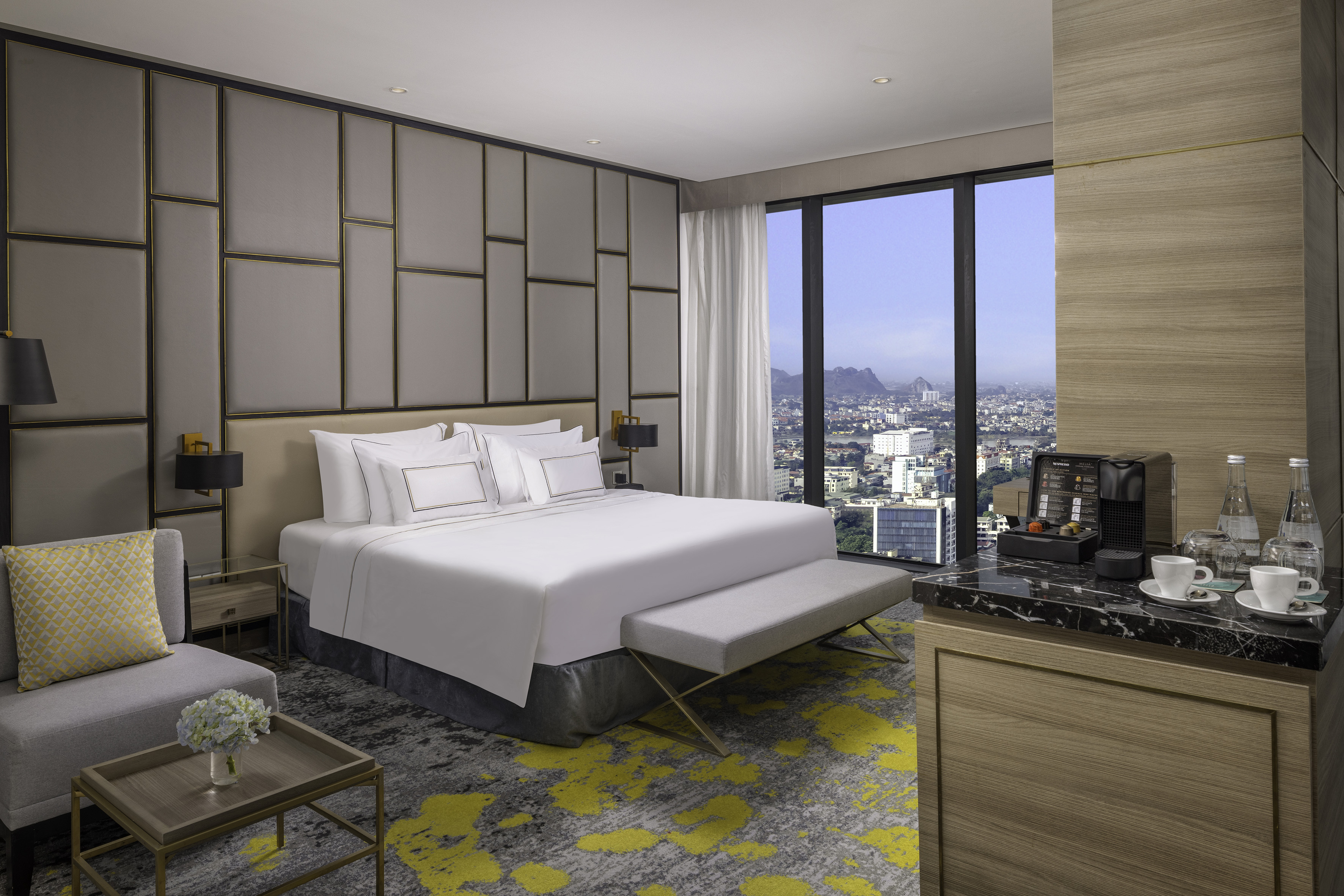 Modern hotel suite with king bed, large window city view, coffee station.
