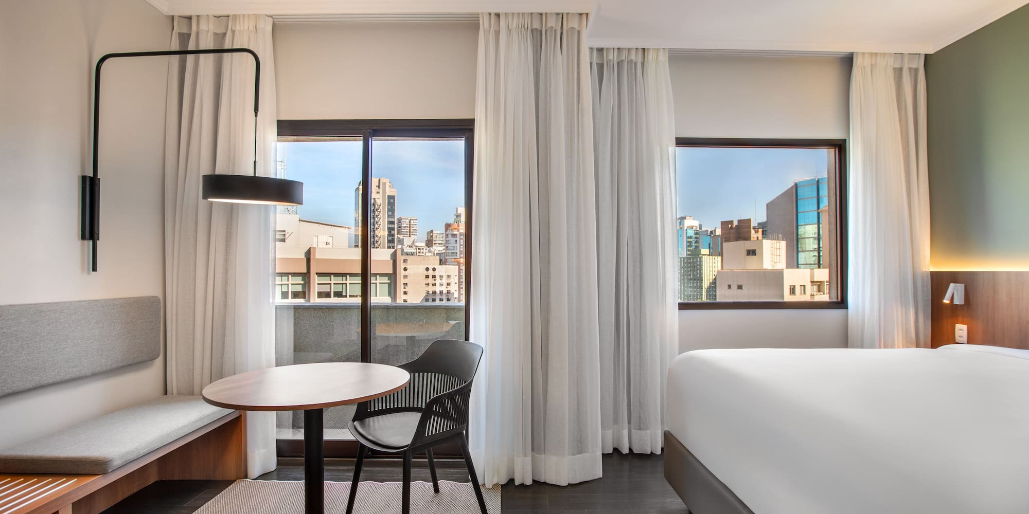 Bright hotel room with bed, table, seating, and urban city view.