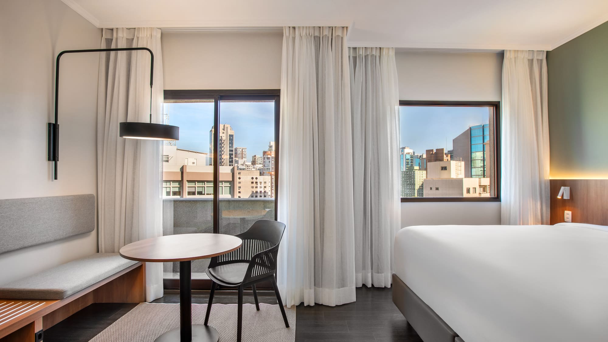 Bright hotel room with bed, table, seating, and urban city view.