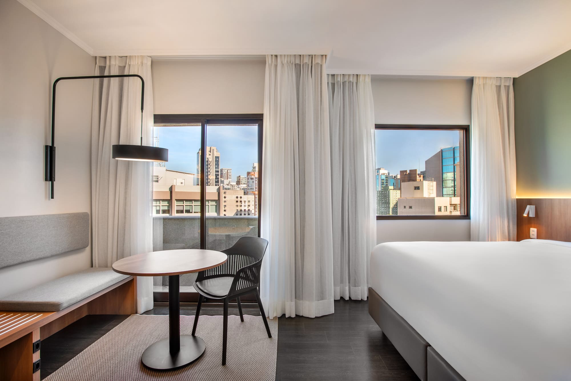 Bright hotel room with bed, table, seating, and urban city view.
