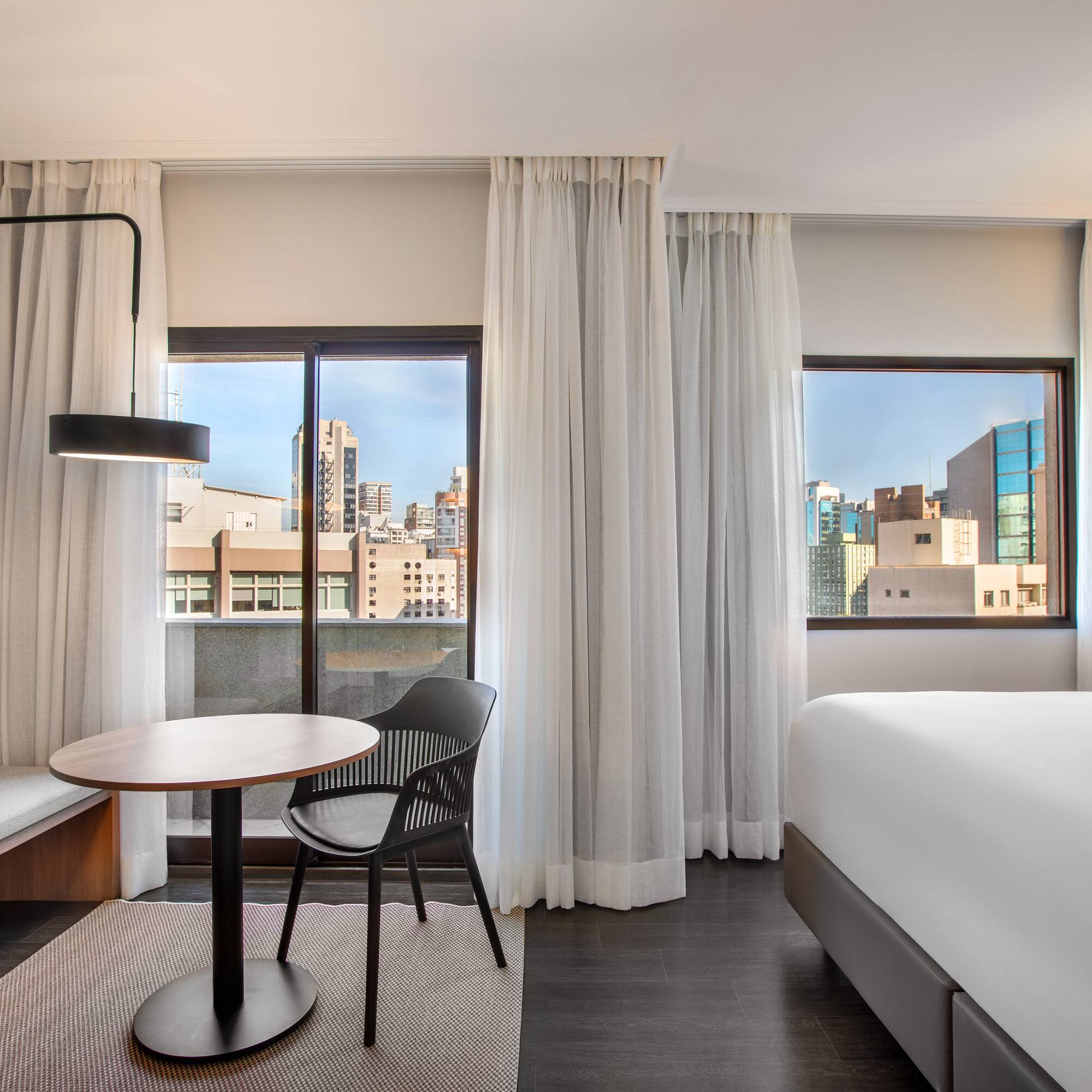 Bright hotel room with bed, table, seating, and urban city view.
