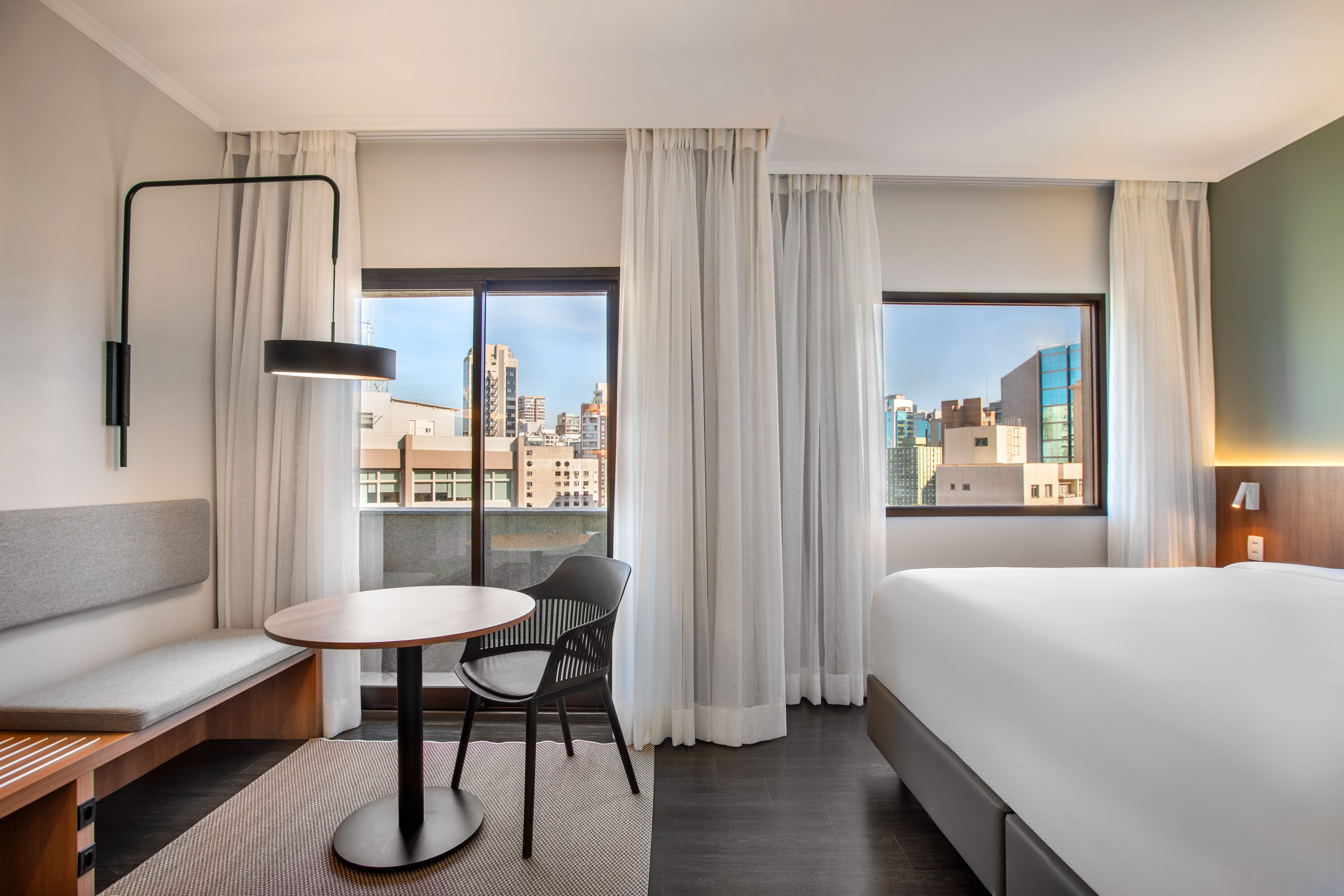 Bright hotel room with bed, table, seating, and urban city view.
