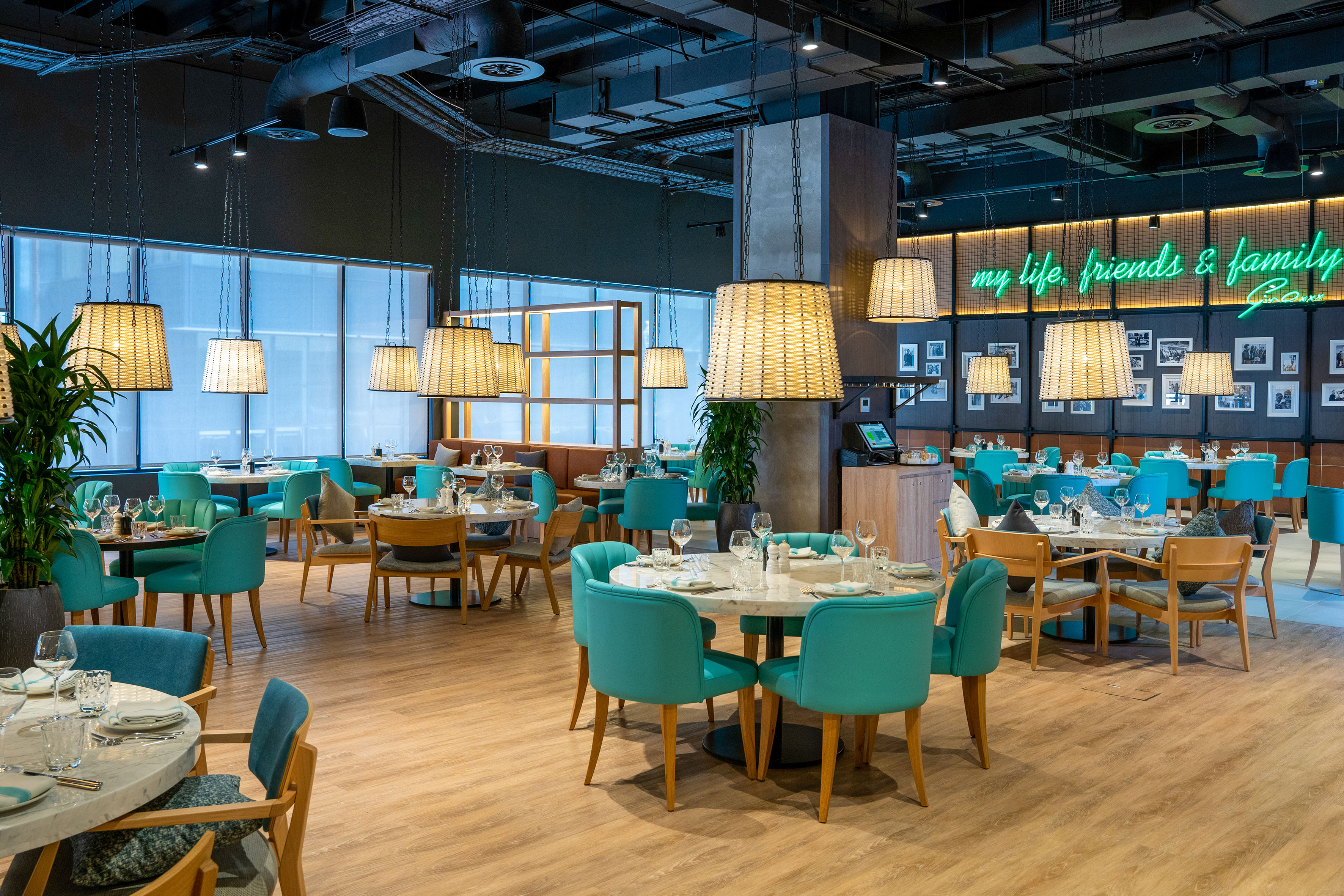 Modern restaurant dining area with teal chairs, marble tables, woven lights, neon sign.