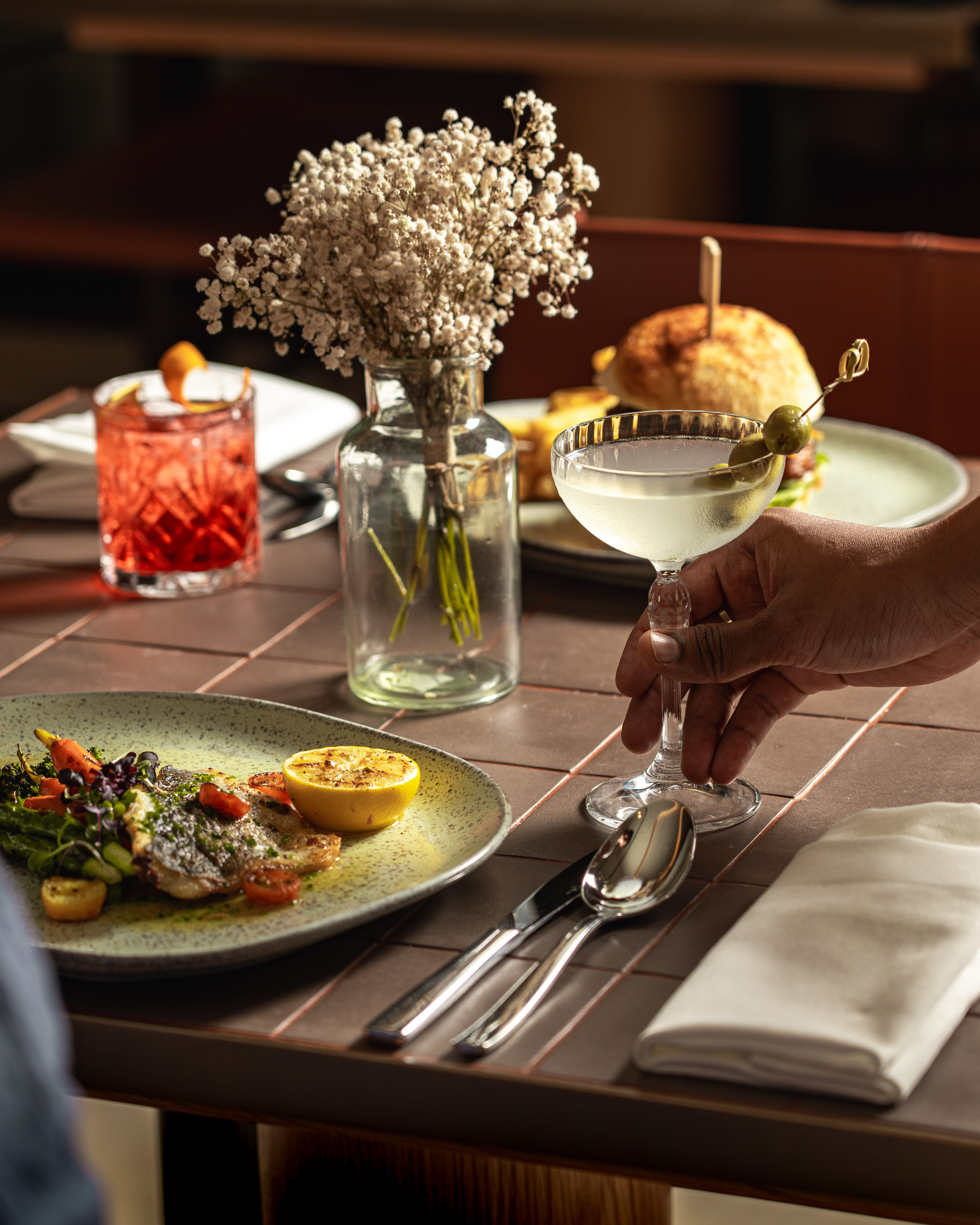 Hand holding martini at restaurant table with grilled fish, cocktail, burger.