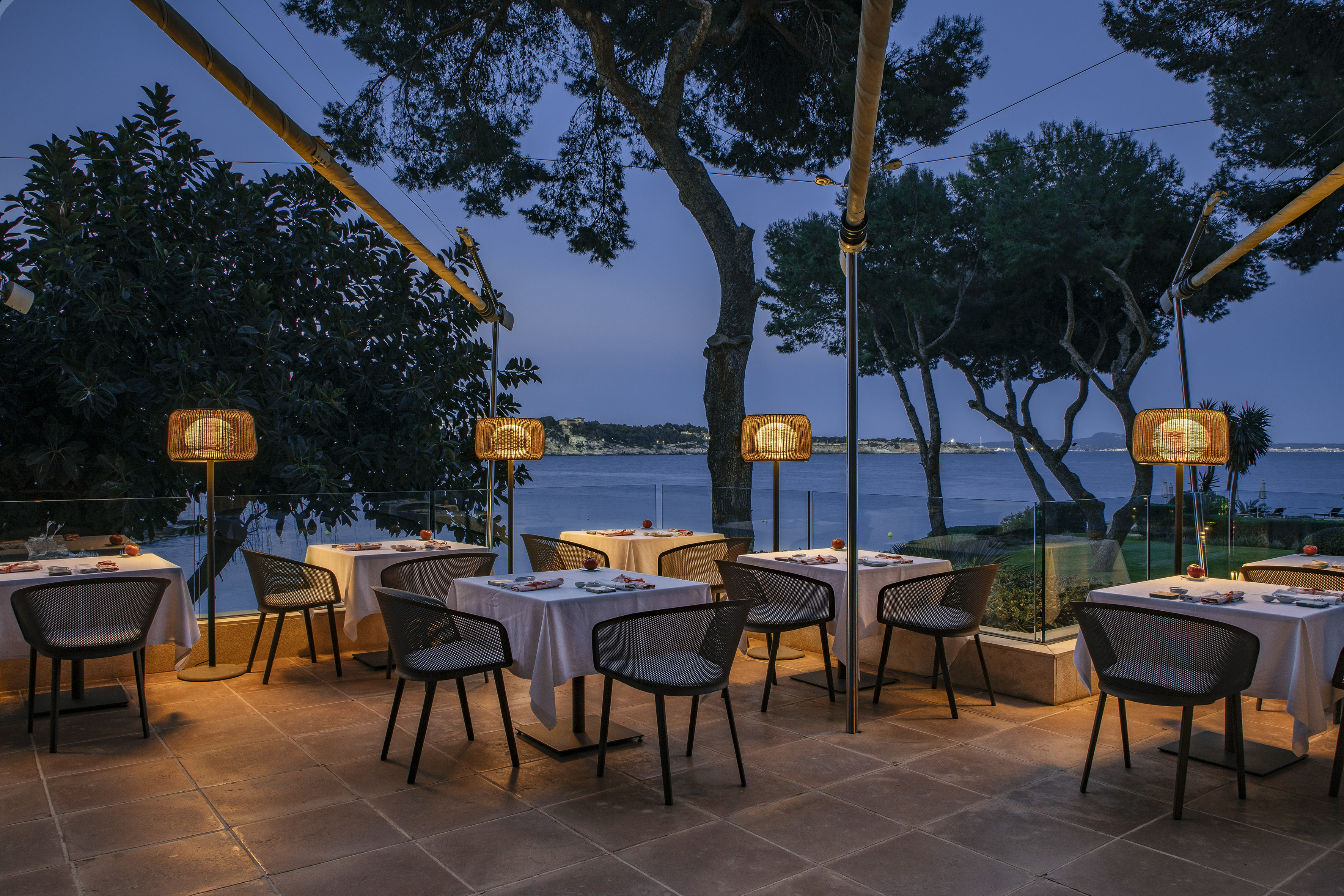 Outdoor restaurant terrace with set tables, glowing lamps, and an ocean view at dusk.
