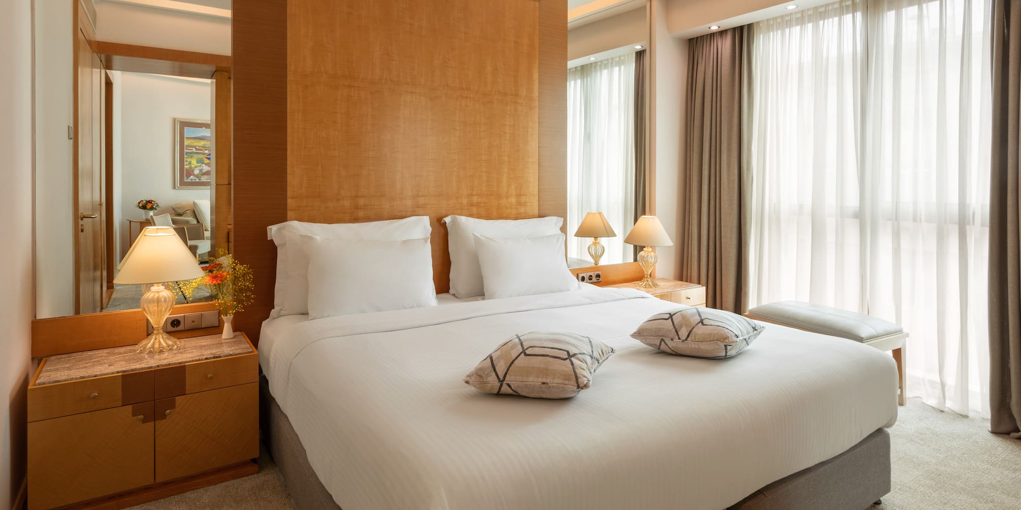Luxury hotel bedroom with large bed, wooden headboard, and bright window.