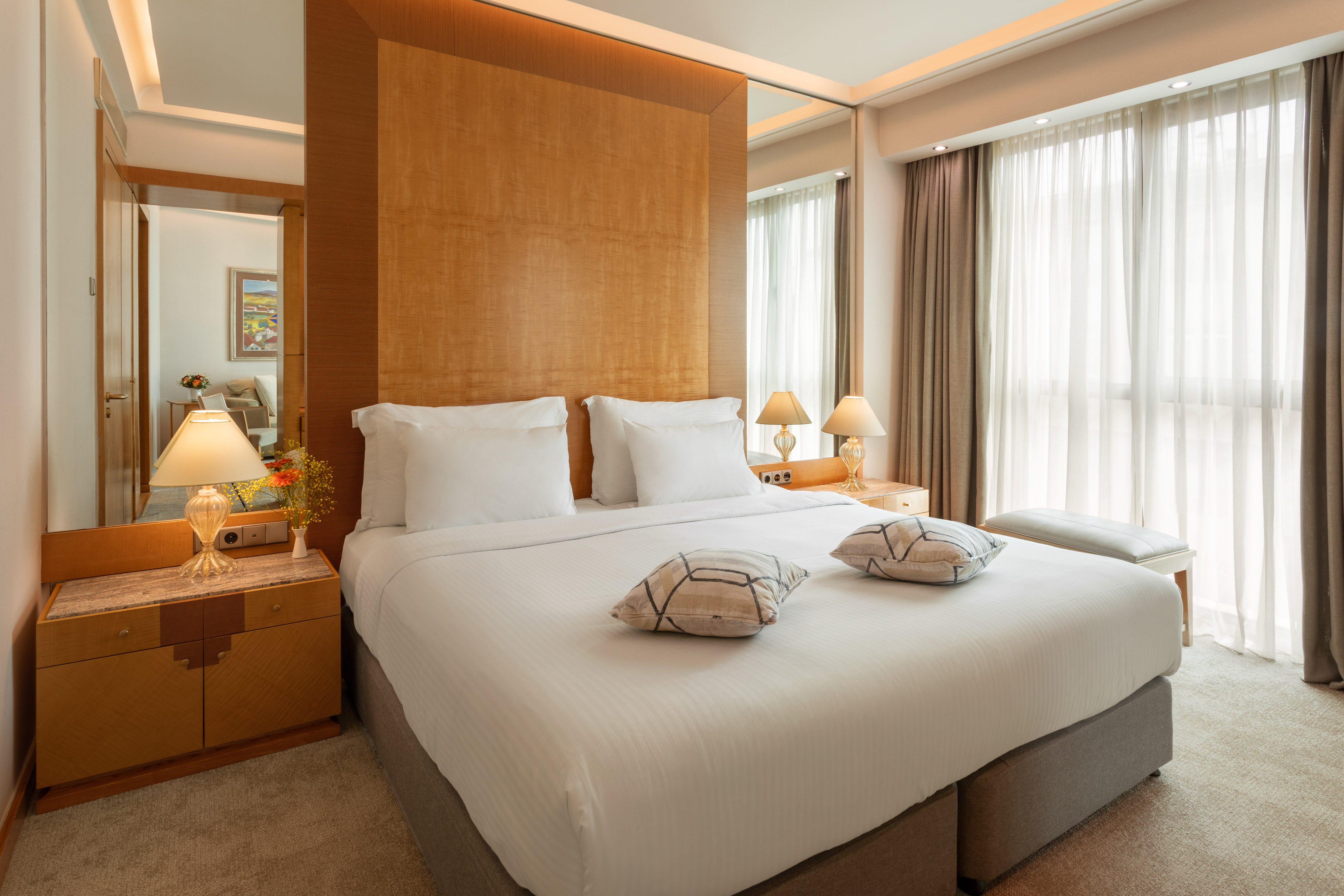 Luxury hotel bedroom with large bed, wooden headboard, and bright window.