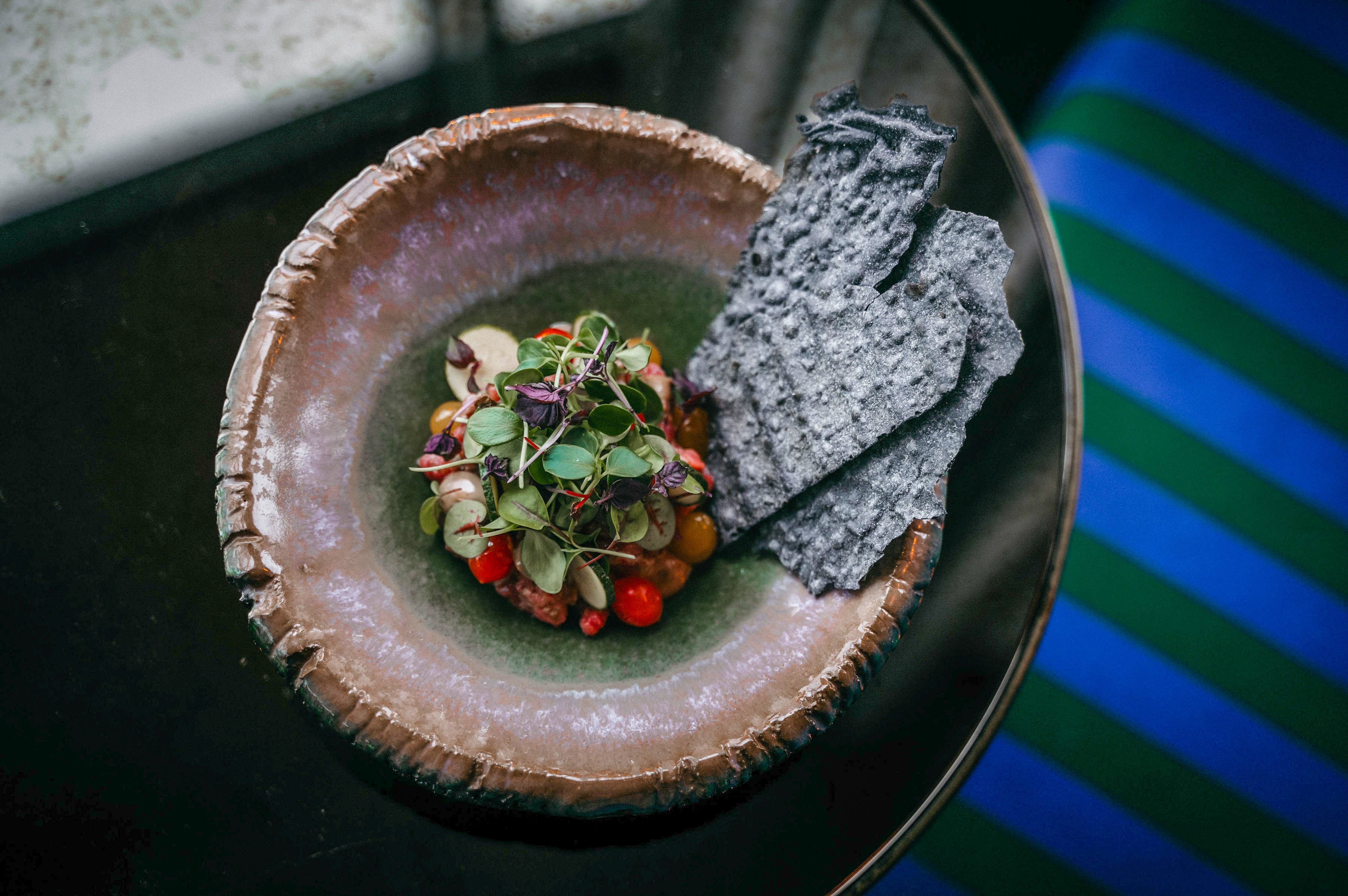 Gourmet tartare with microgreens, tomatoes, and charcoal crisps in rustic bowl.