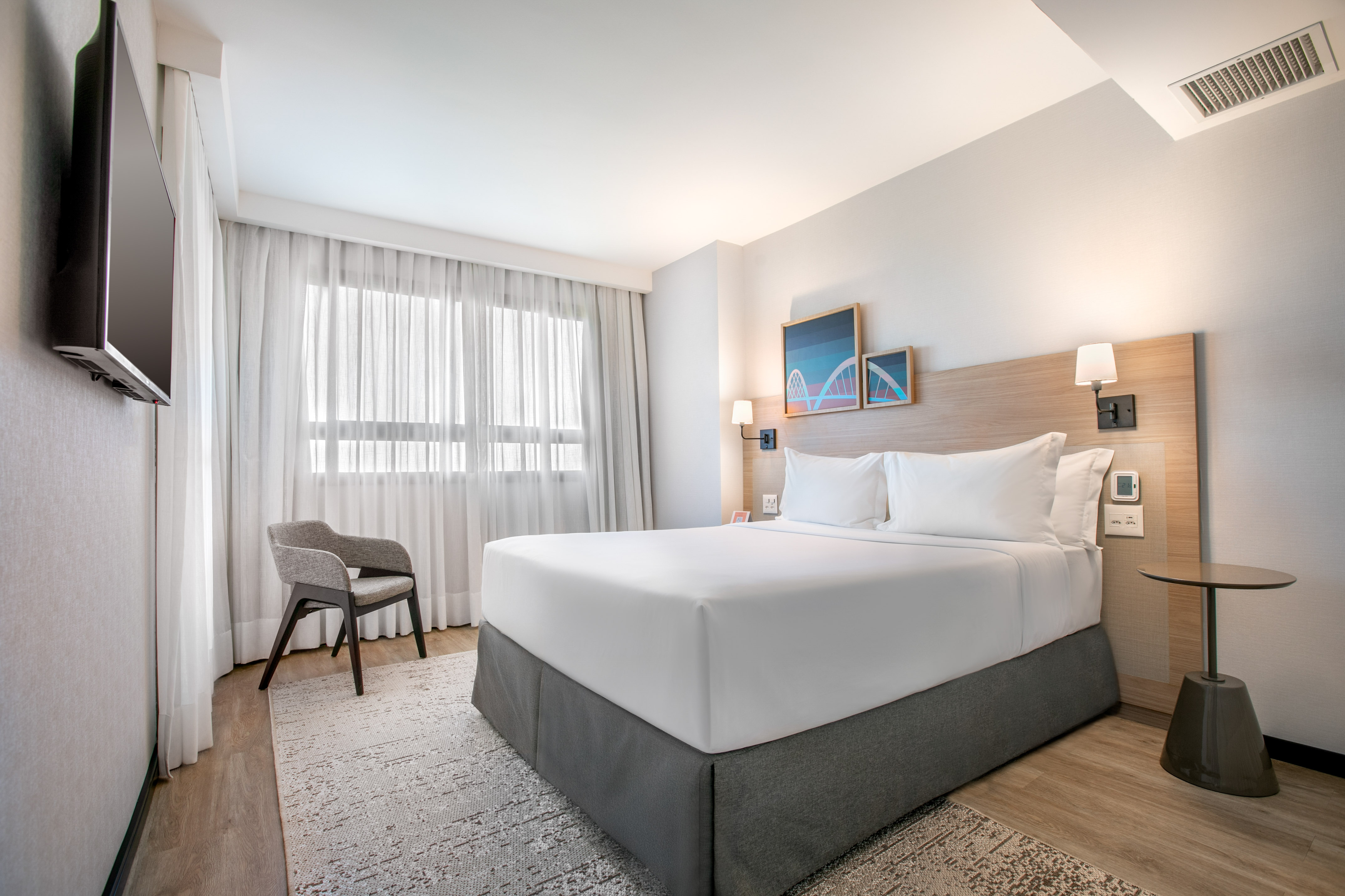 Modern hotel bedroom with white bed, wall art, armchair, and bright window.
