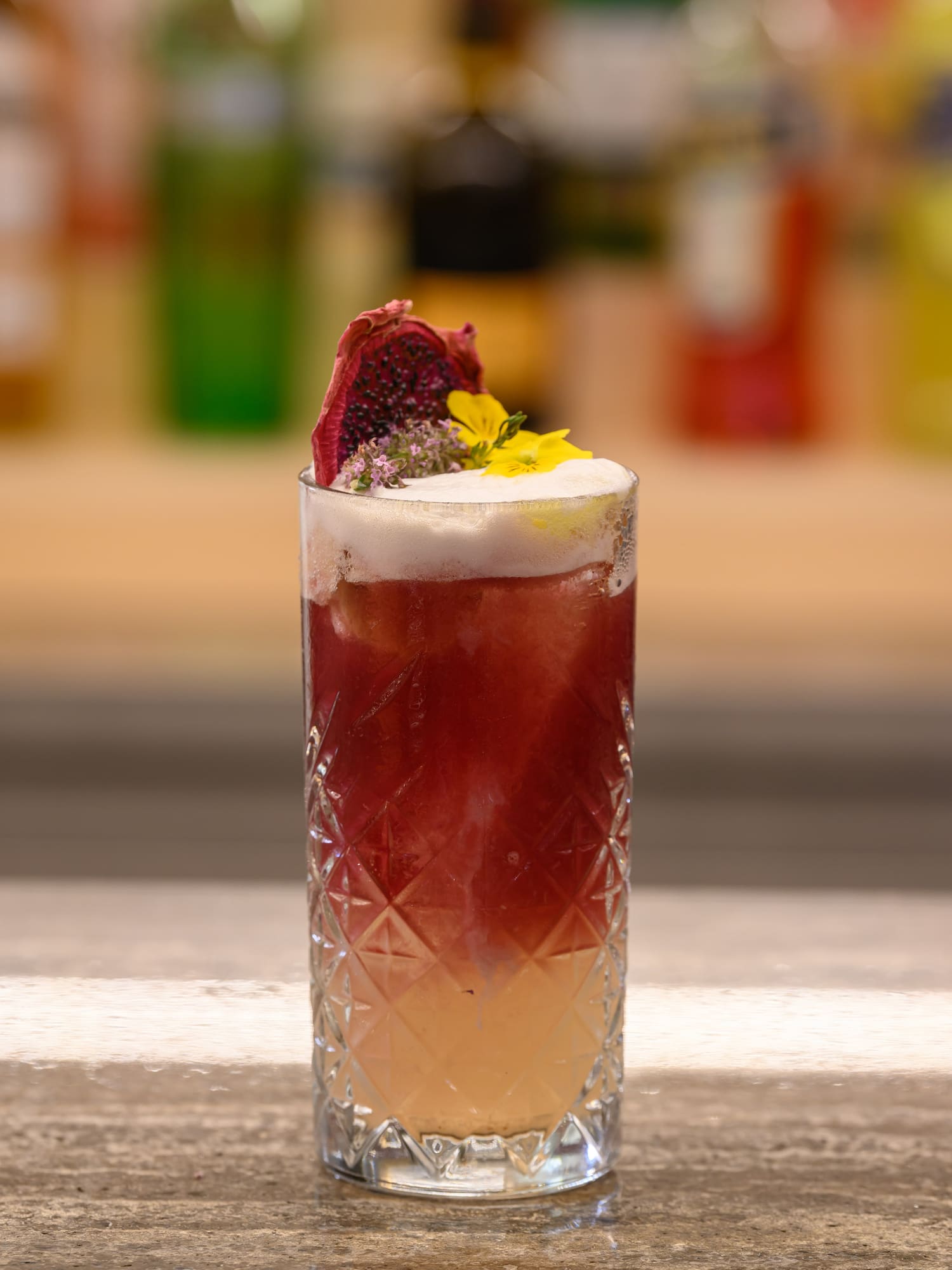 Layered red cocktail in textured glass, garnished with dried dragon fruit and yellow flowers.