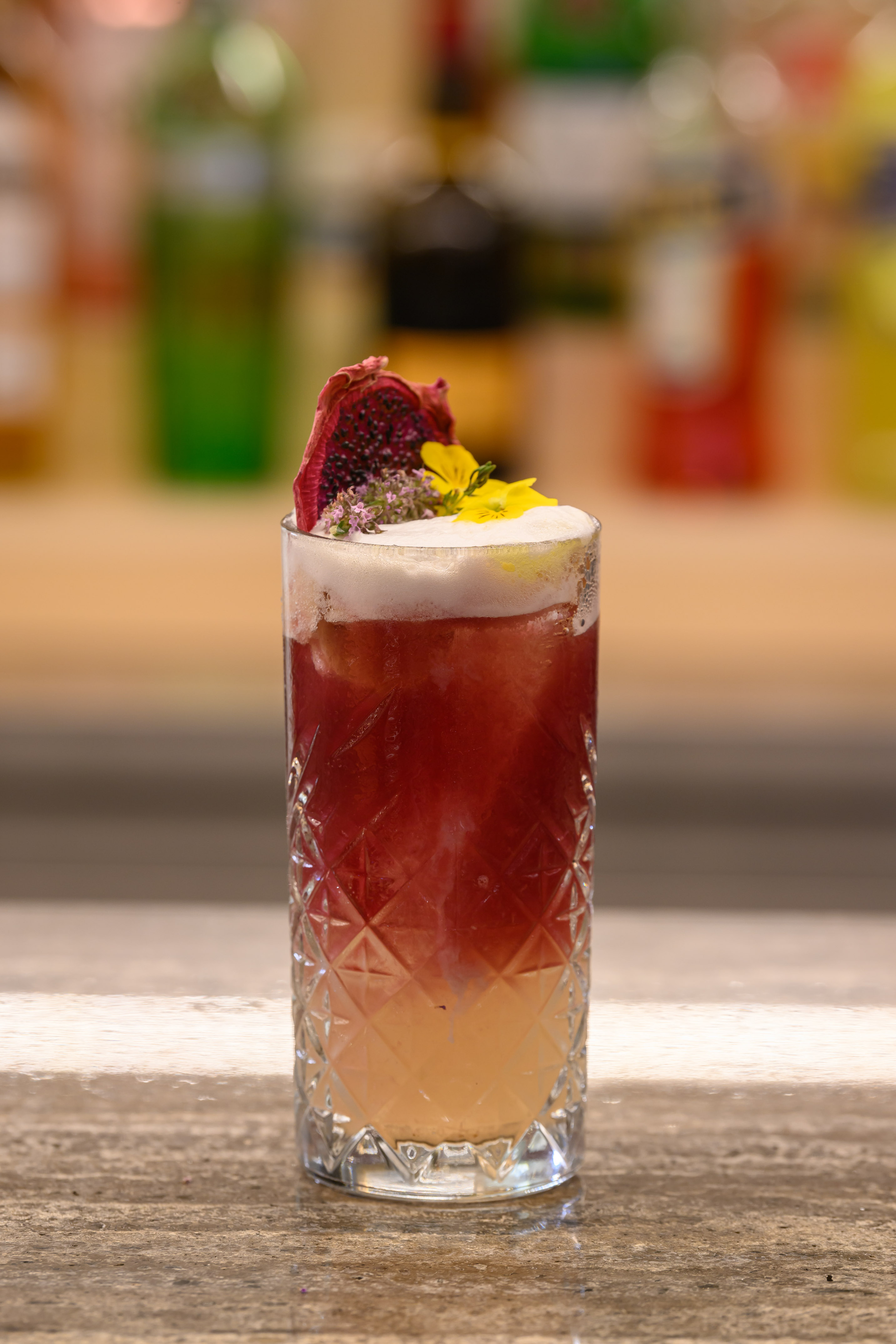 Layered red cocktail in textured glass, garnished with dried dragon fruit and yellow flowers.