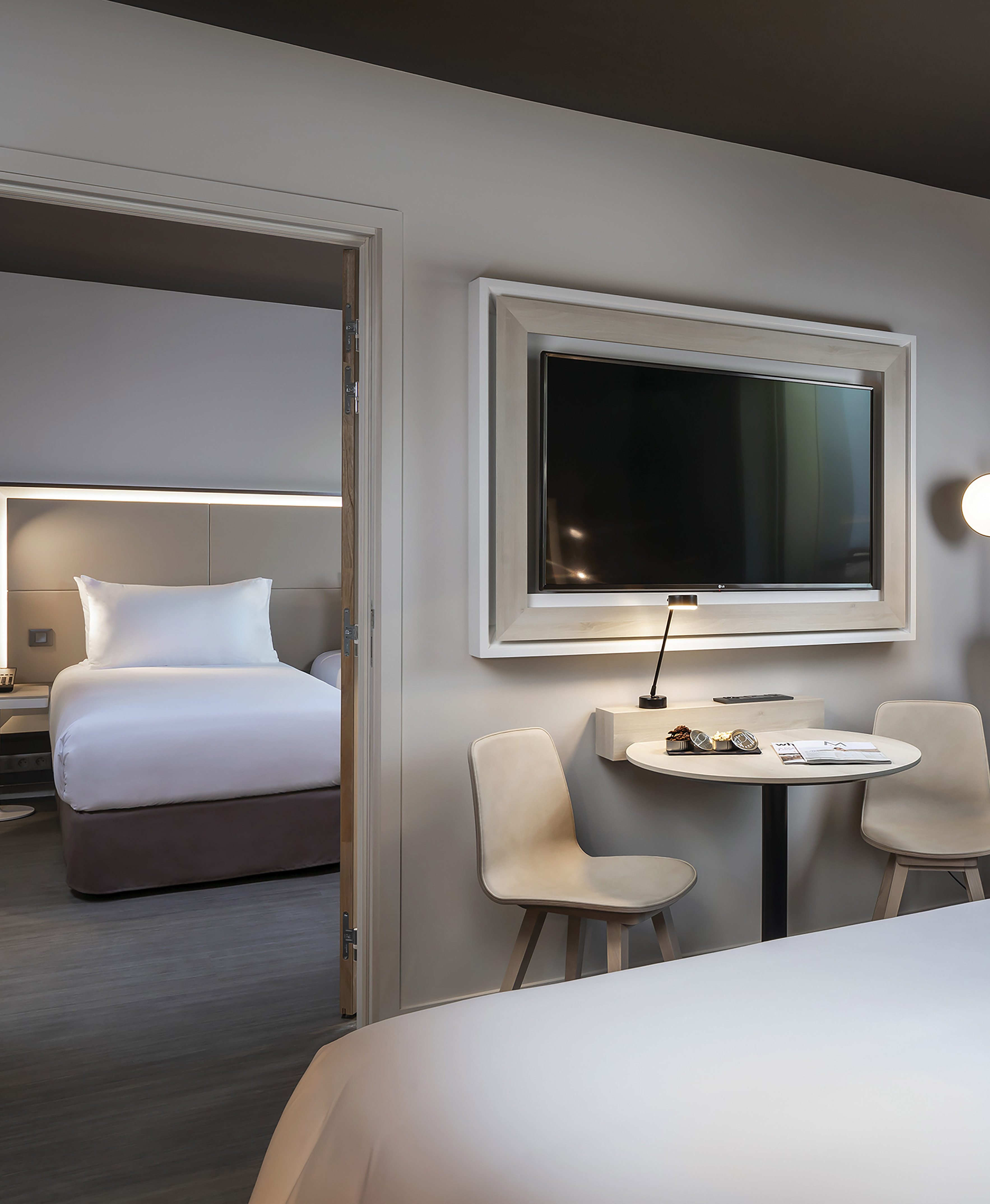 Contemporary hotel room with wall-mounted TV, table, and adjacent bed.