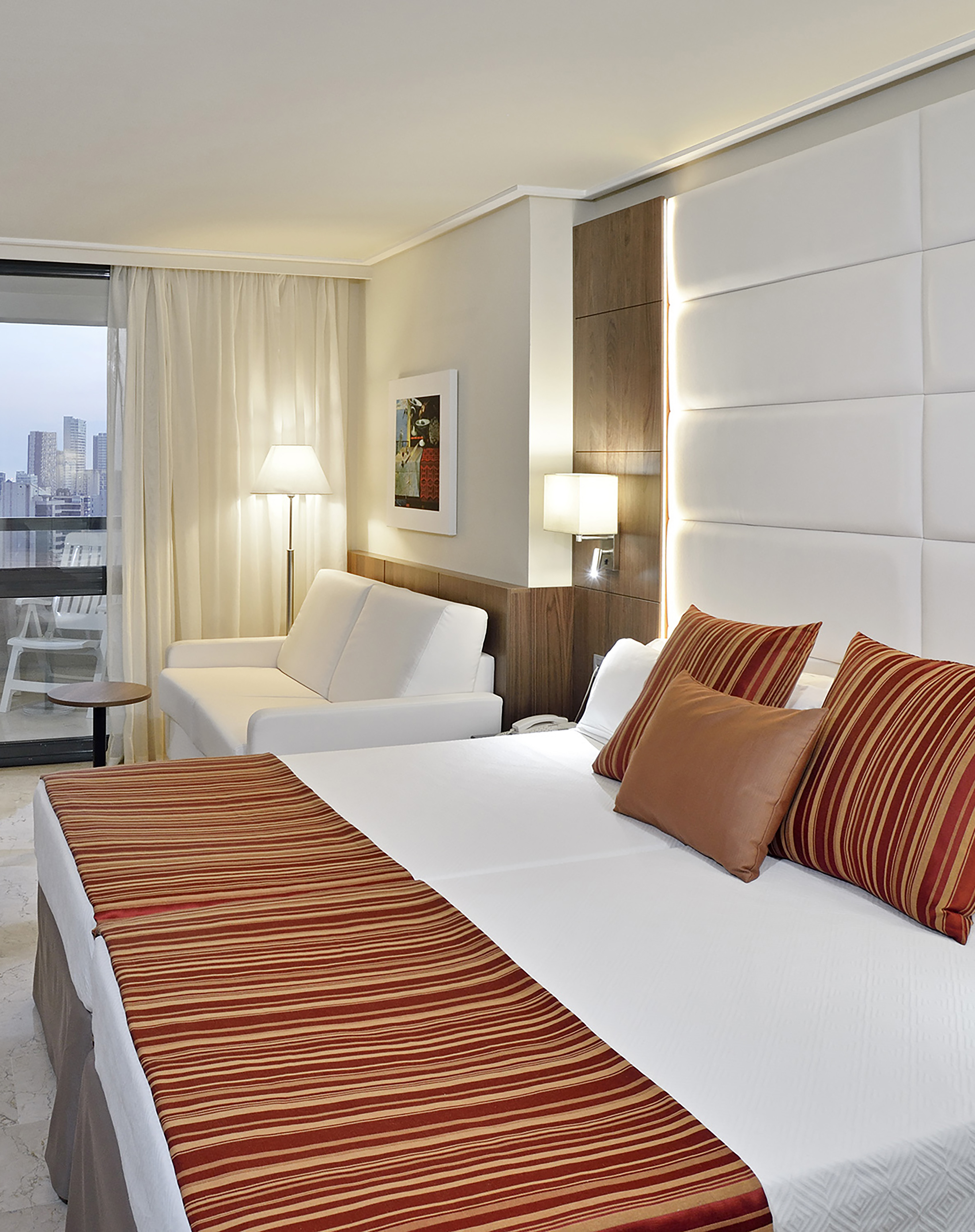 Modern hotel room with king bed, white sofa, city view balcony, and striped bedding.