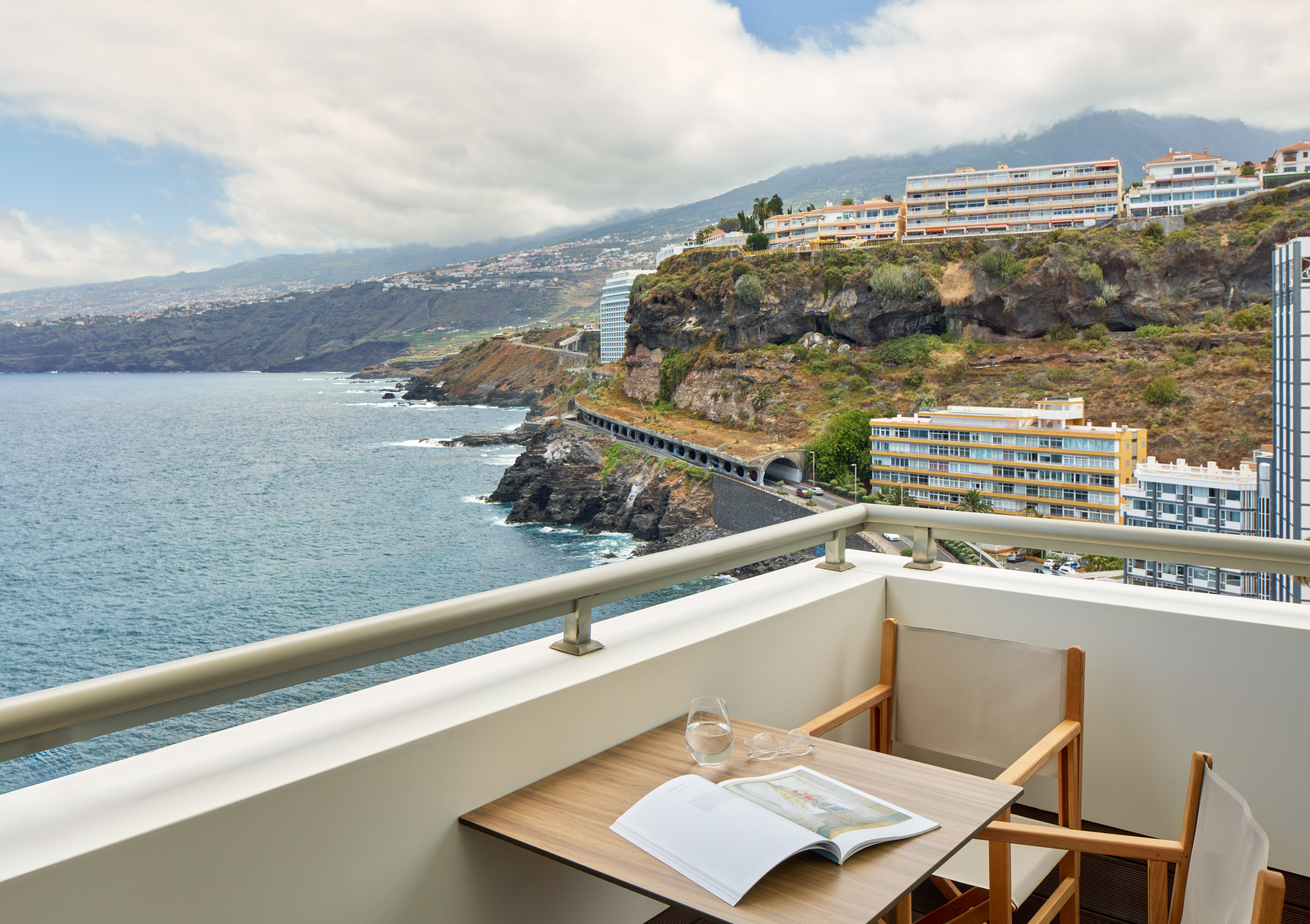 Hotel balcony ocean view with book, coastal town on cliffs, mountains.