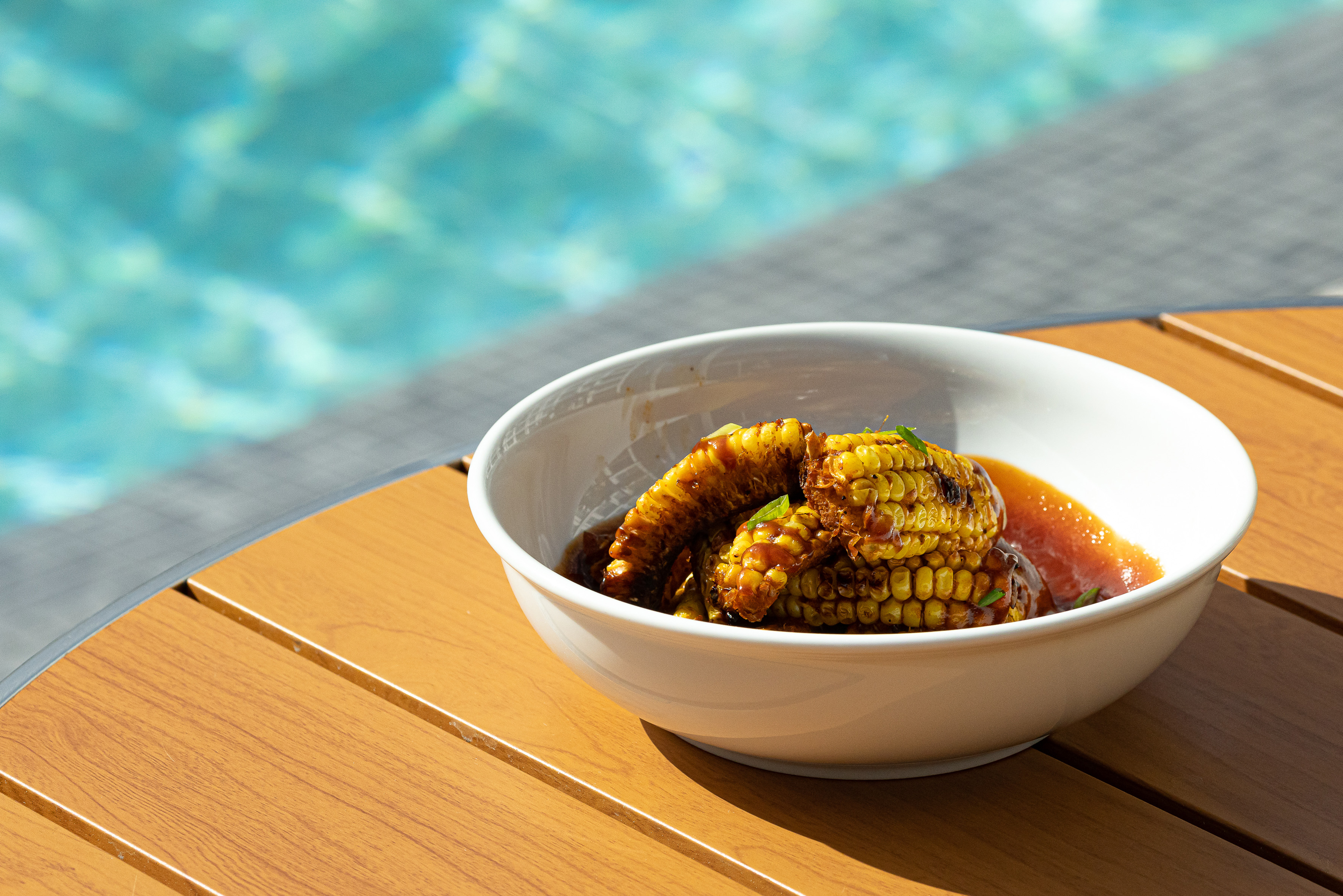 Grilled corn in sauce in a white bowl on a wooden table beside a blue pool.