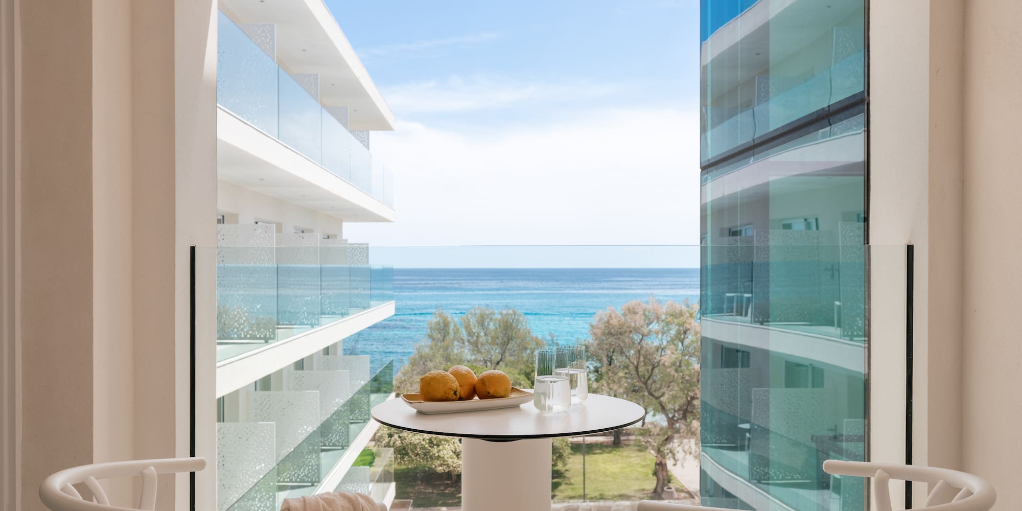 Modern hotel balcony with sea view, white table with lemons, and chairs.