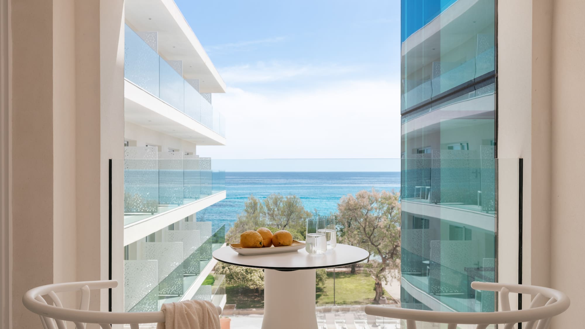 Modern hotel balcony with sea view, white table with lemons, and chairs.