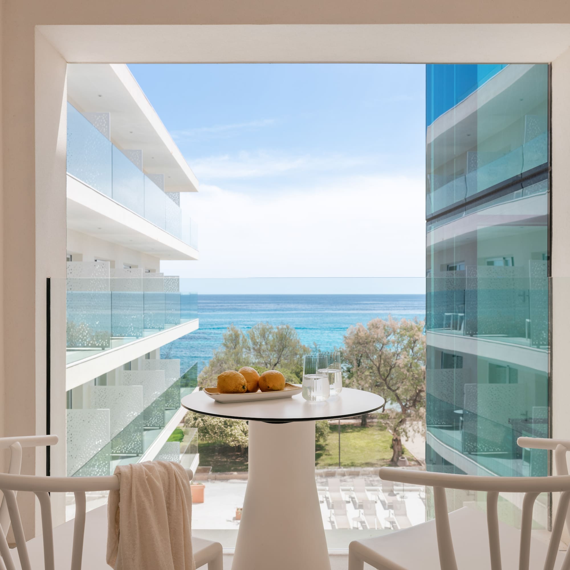 Modern hotel balcony with sea view, white table with lemons, and chairs.