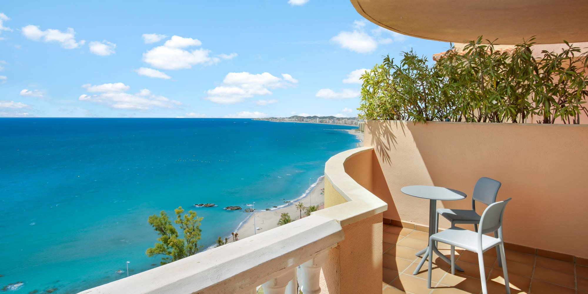 Resort balcony with blue ocean, sandy beach, and distant coastal city view.
