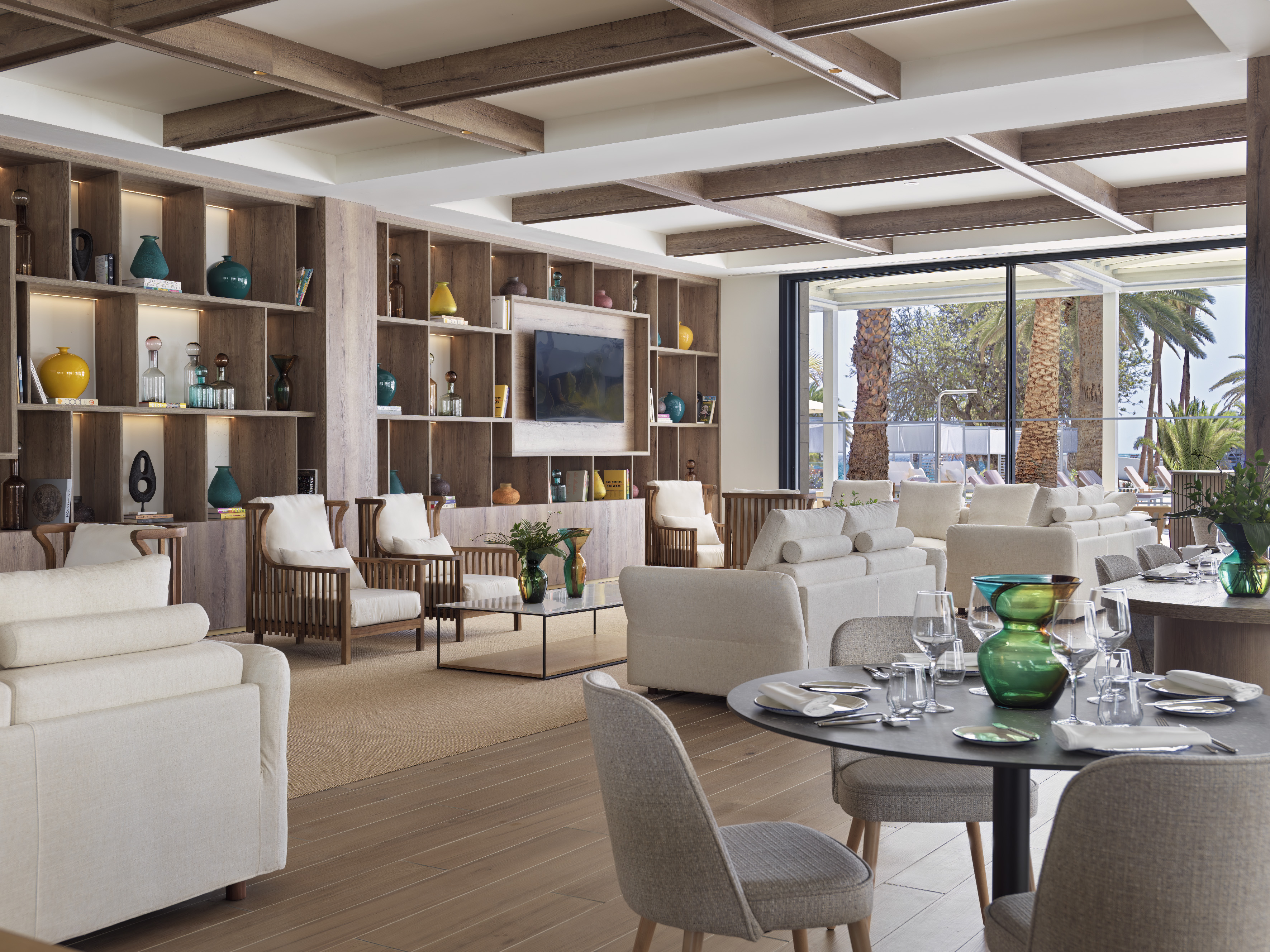 Modern resort lounge with wood shelves, soft seating, and outdoor palm view.
