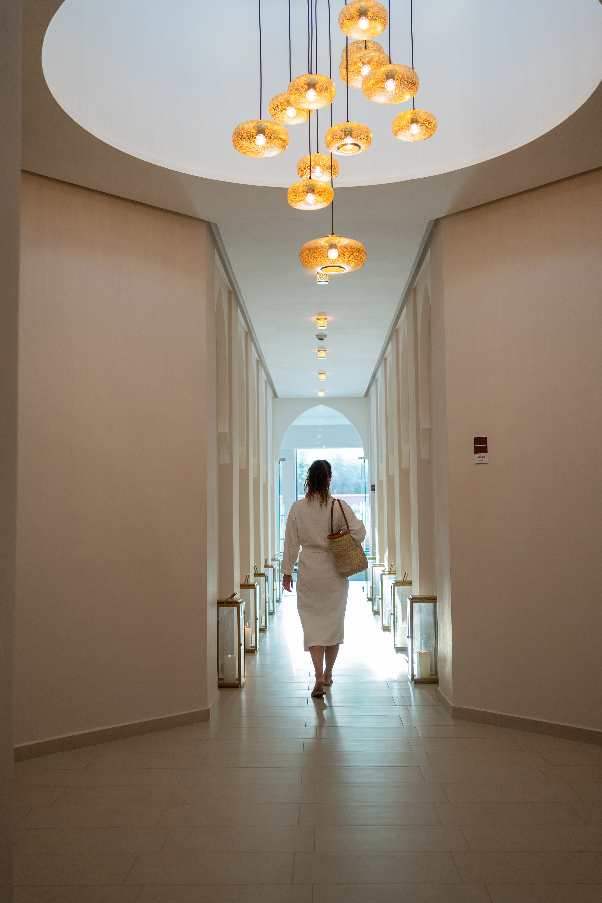 Woman in white robe walks elegant hotel spa corridor with glowing lights.