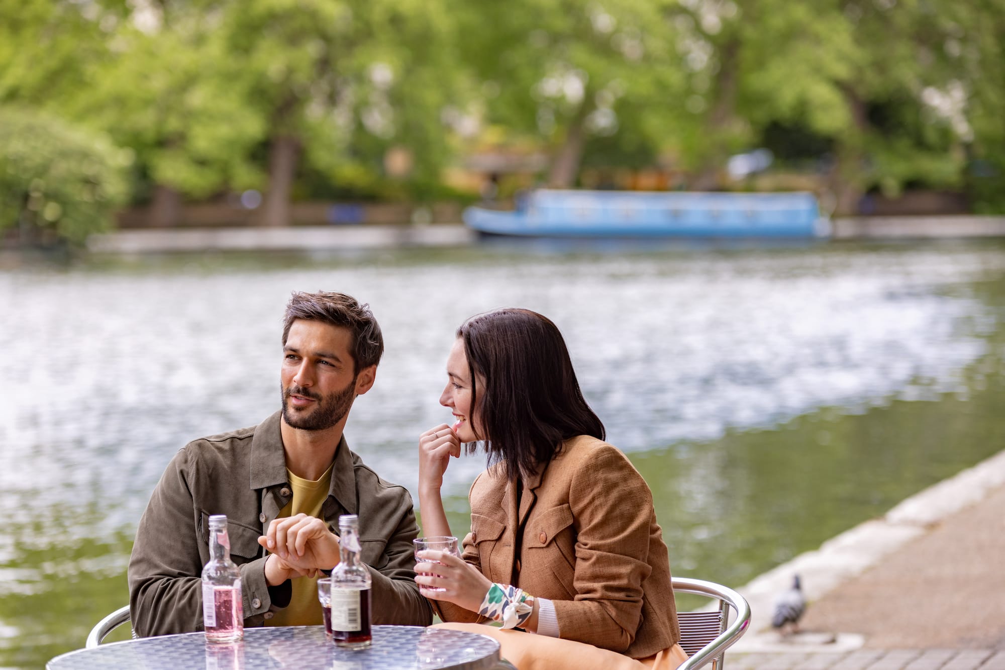 Pareja conversando en terraza fluvial con bebidas y barco al fondo.