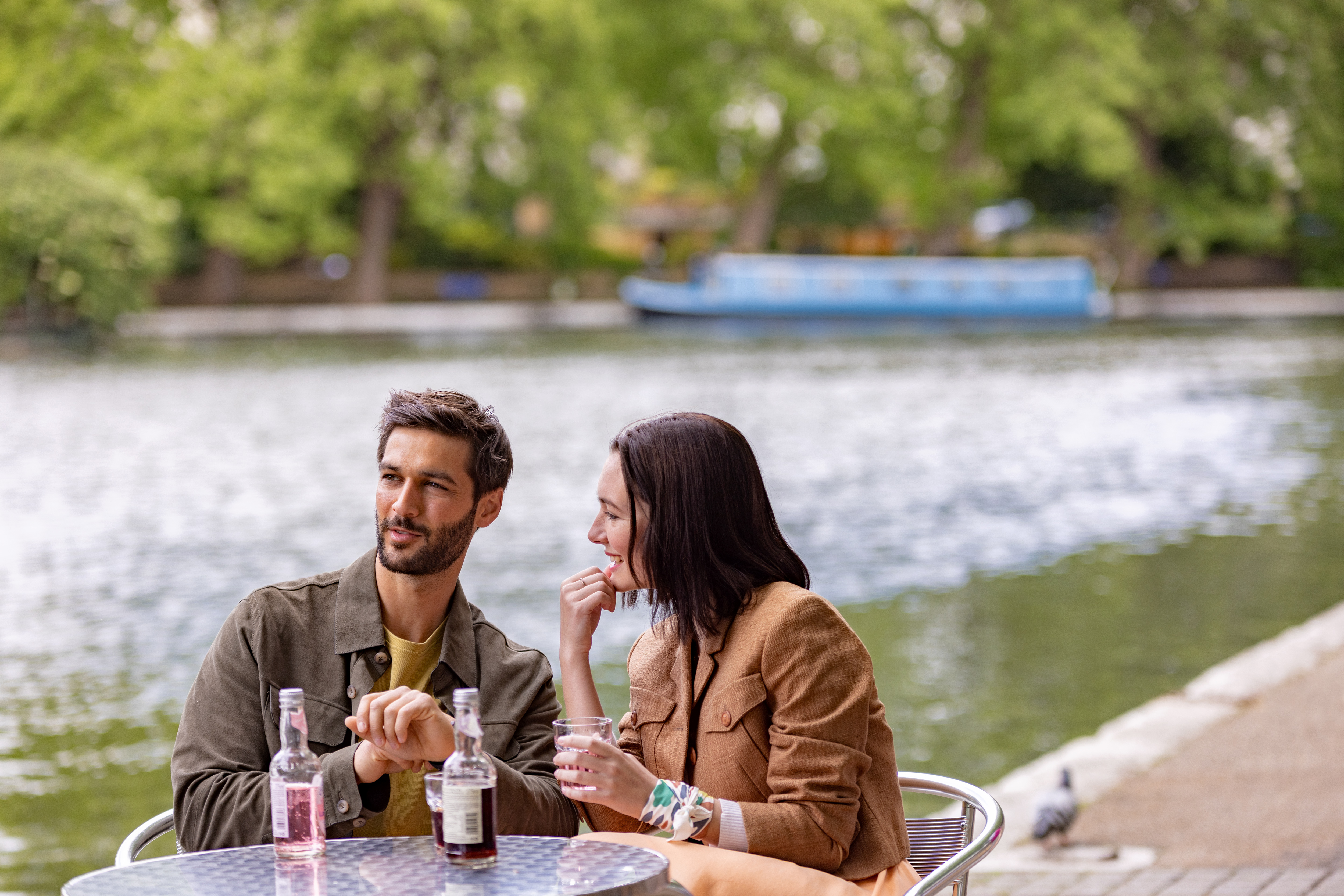 Pareja conversando en terraza fluvial con bebidas y barco al fondo.