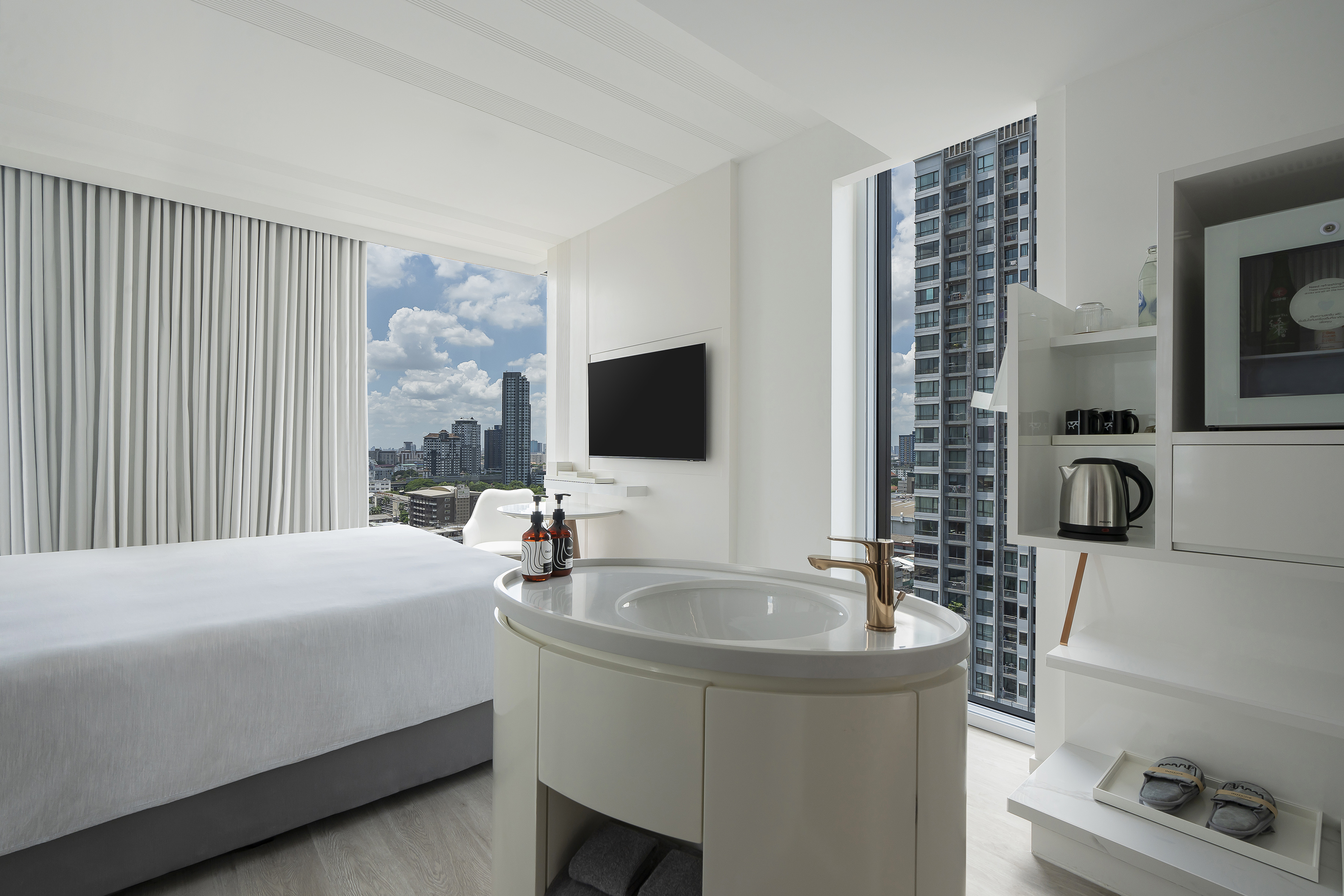 Modern hotel room with city view, white bed, minimalist decor, and vanity sink.