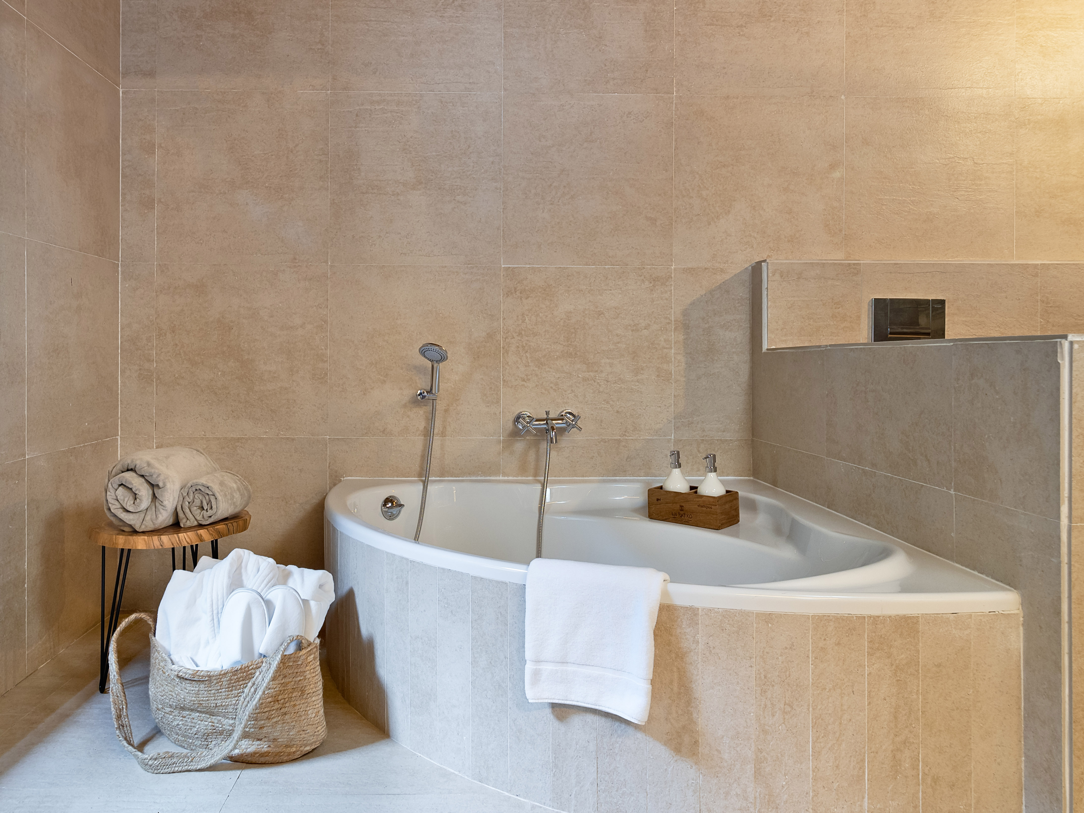 Corner bathtub in a modern bathroom with beige tiles, towels, and toiletries.