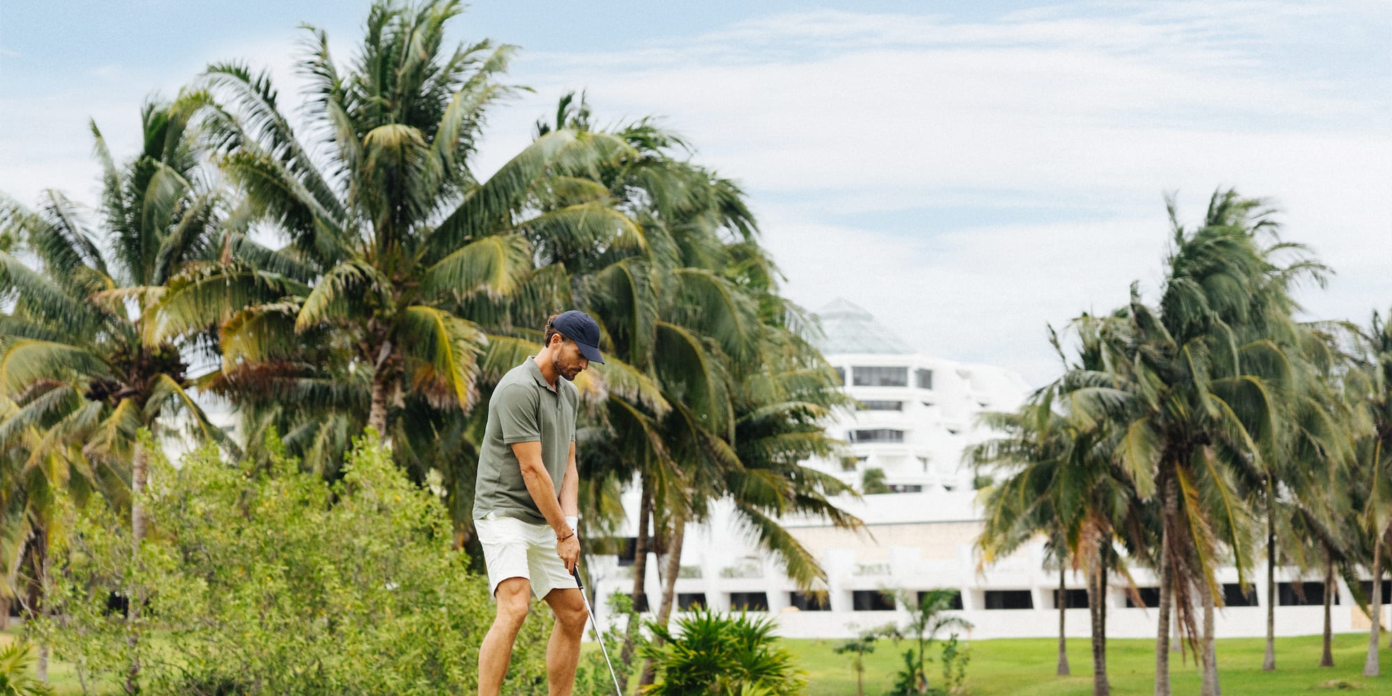 Golfista preparándose para golpear la bola en campo tropical con palmeras y resort.