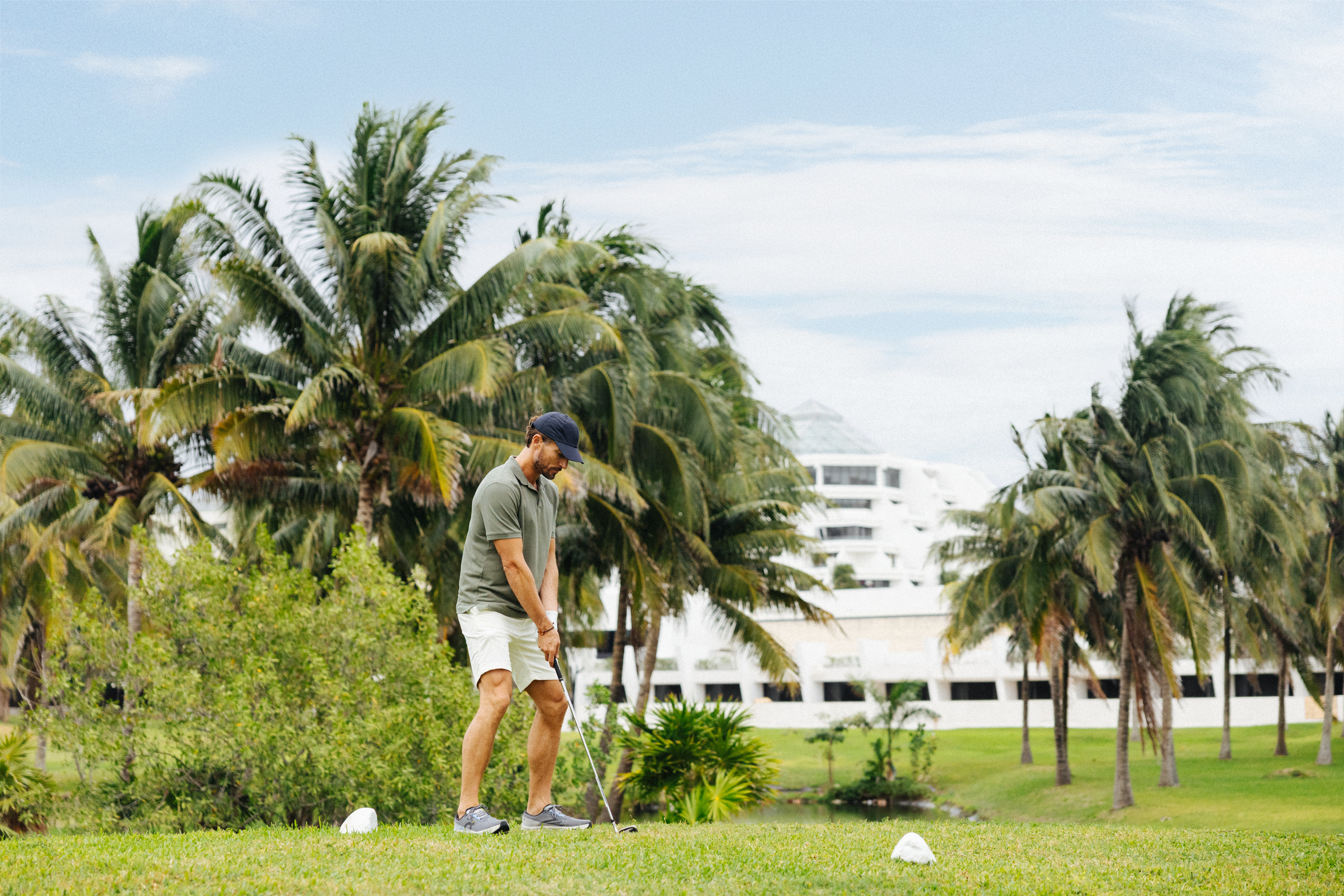 Golfista preparándose para golpear la bola en campo tropical con palmeras y resort.