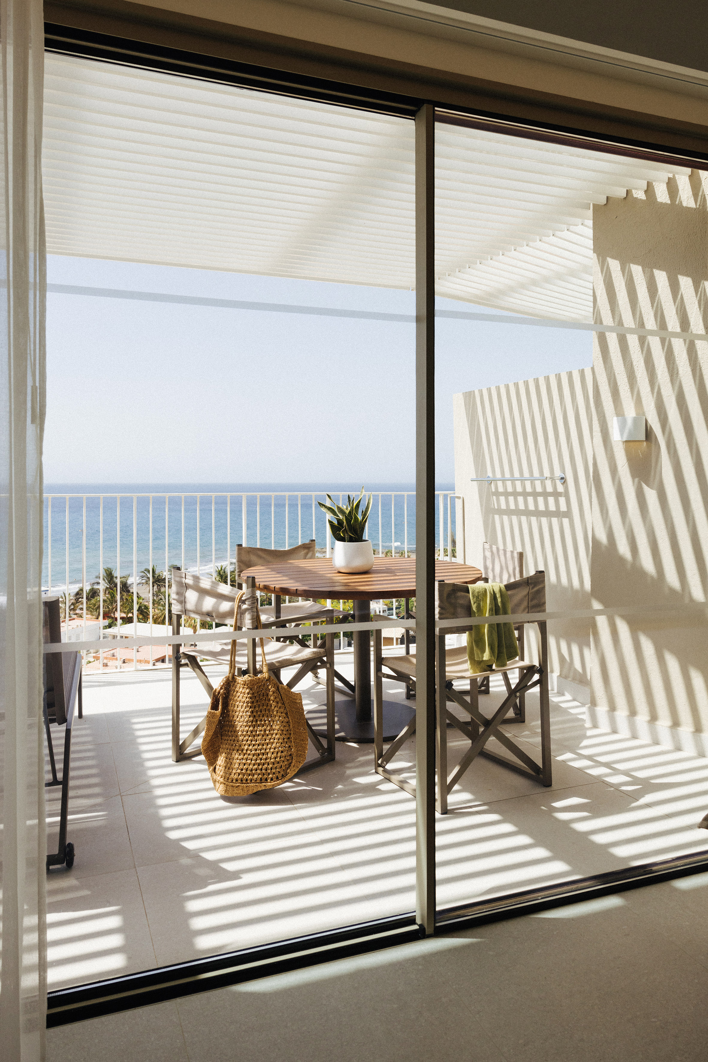 Oceanfront balcony with outdoor dining set, potted plant, and strong sunlit shadows.