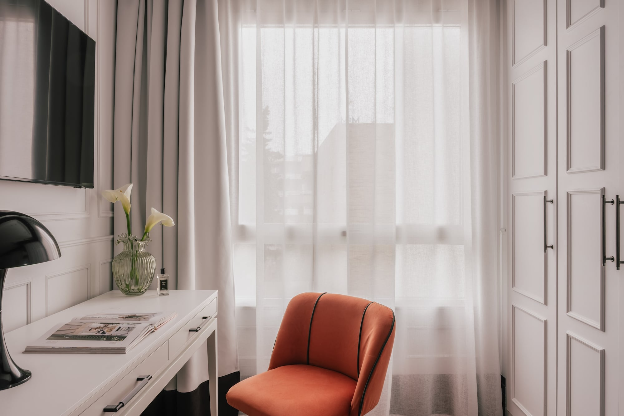 Modern room with white desk, orange velvet chair, TV, and bright window.