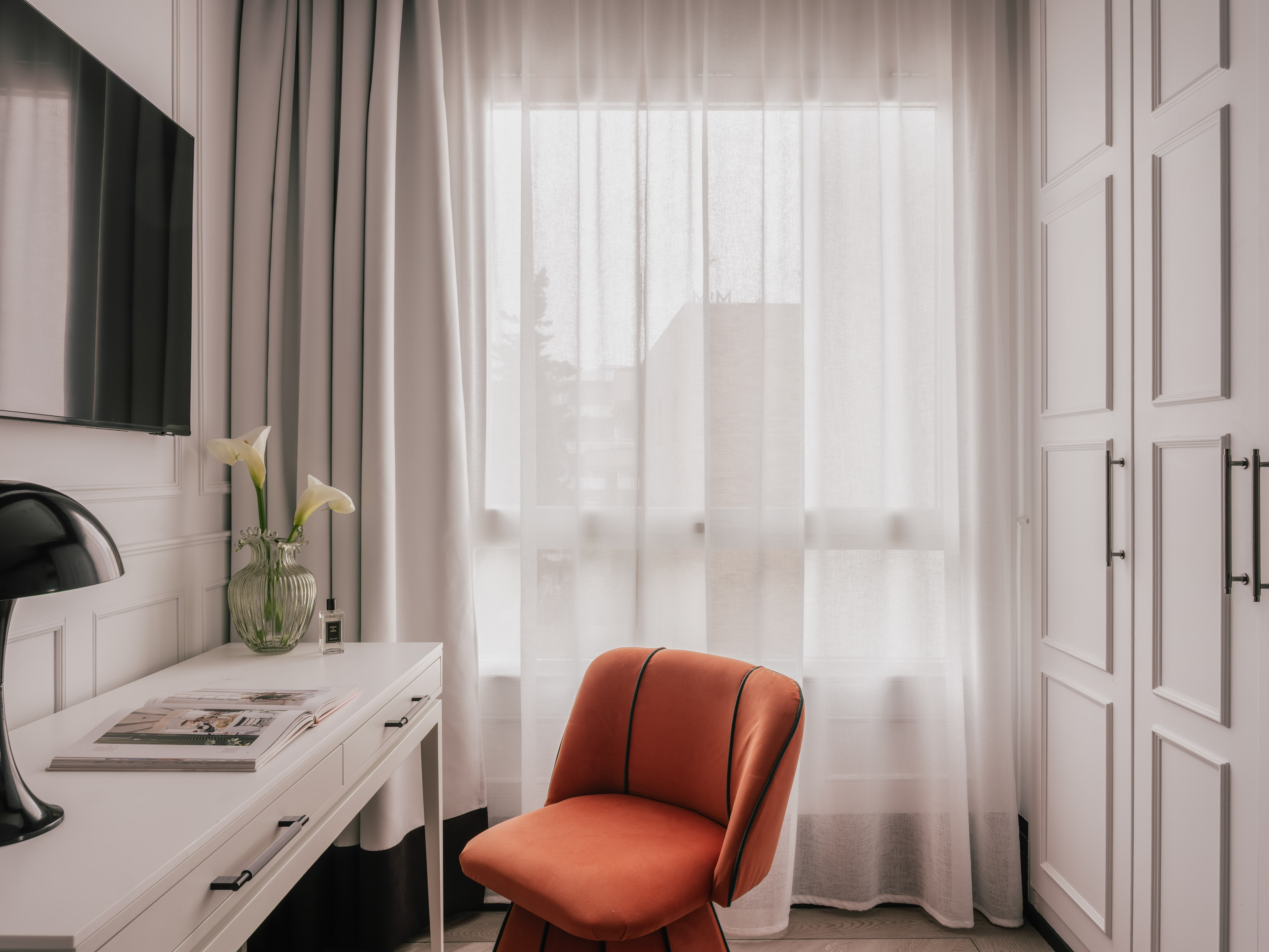 Modern room with white desk, orange velvet chair, TV, and bright window.