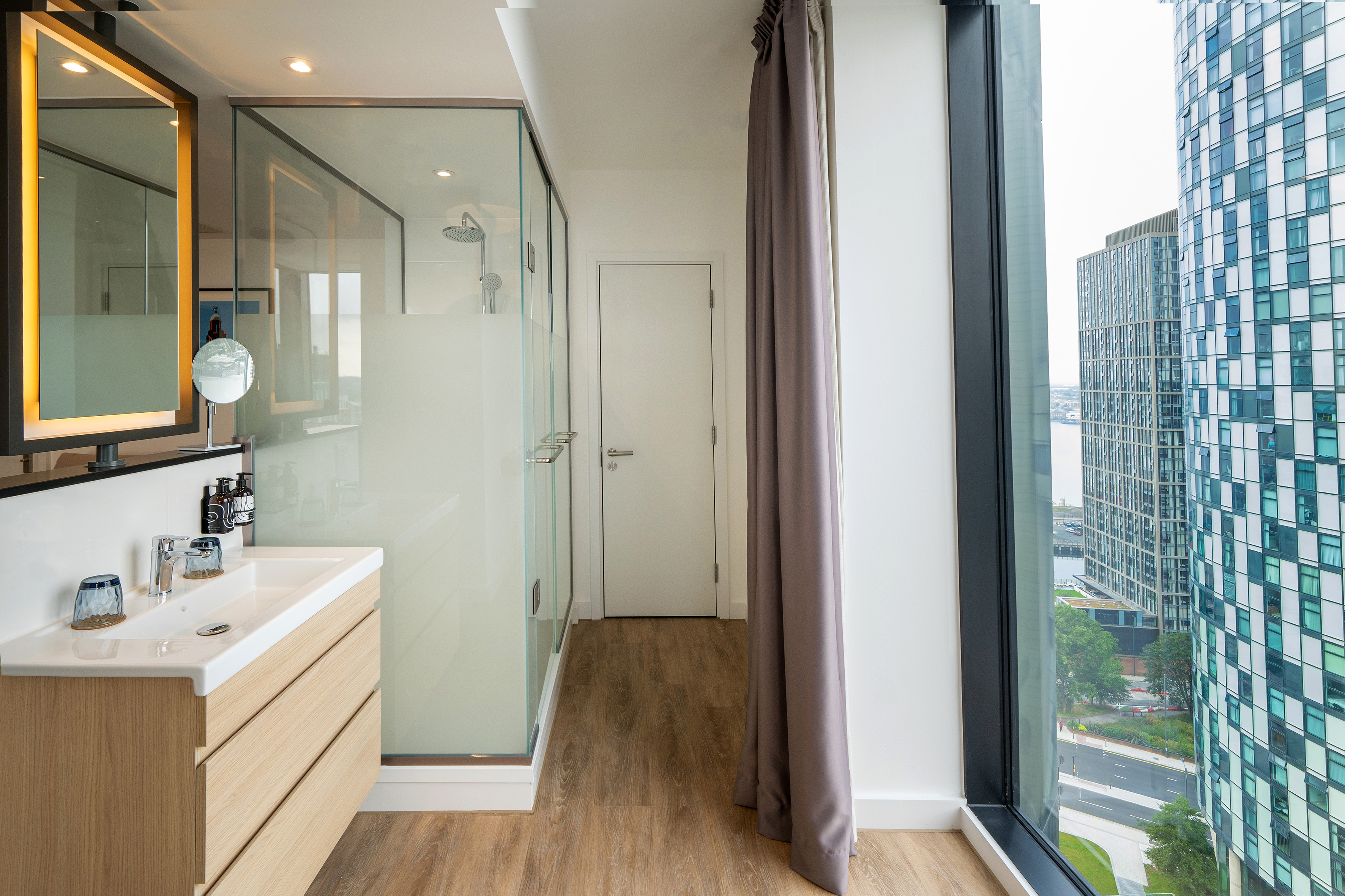 Contemporary bathroom with wood vanity, glass shower, and city view.