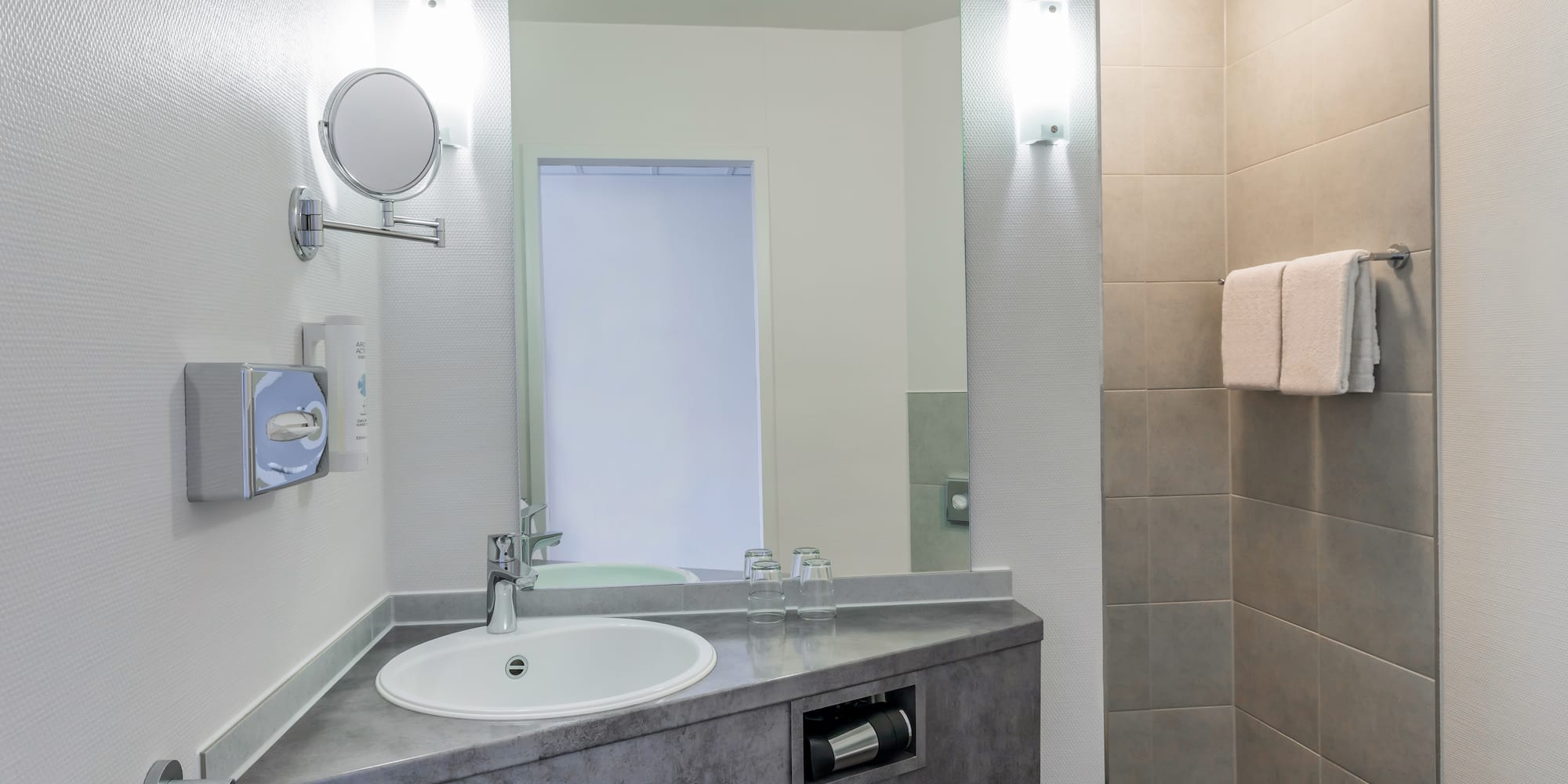 Modern hotel bathroom with sink, large mirror, magnifying mirror, and tiled shower.