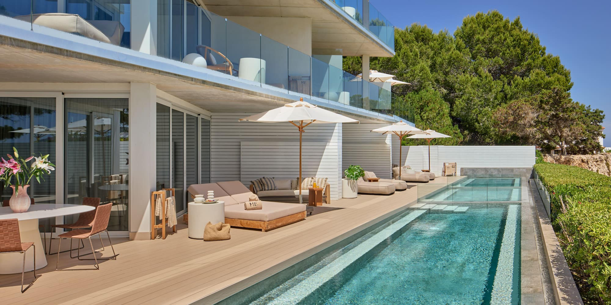 Modern hotel pool deck with sun loungers, umbrellas, balconies, and green trees.