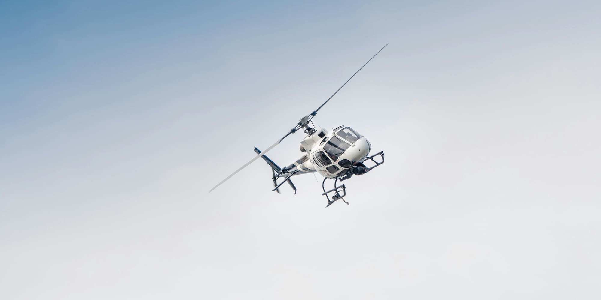 White helicopter with a camera pod flying against a clear, light blue sky.
