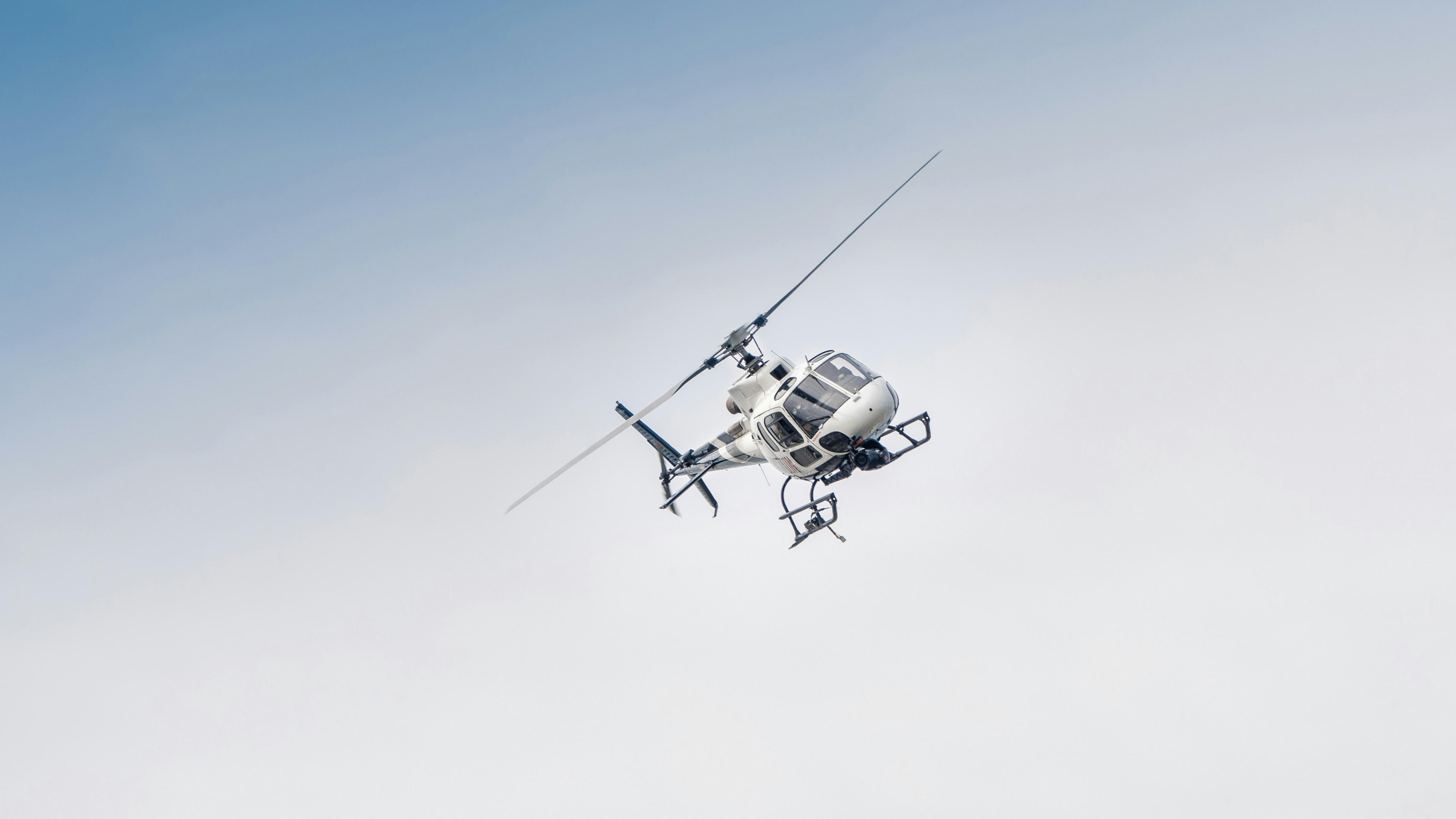 White helicopter with a camera pod flying against a clear, light blue sky.
