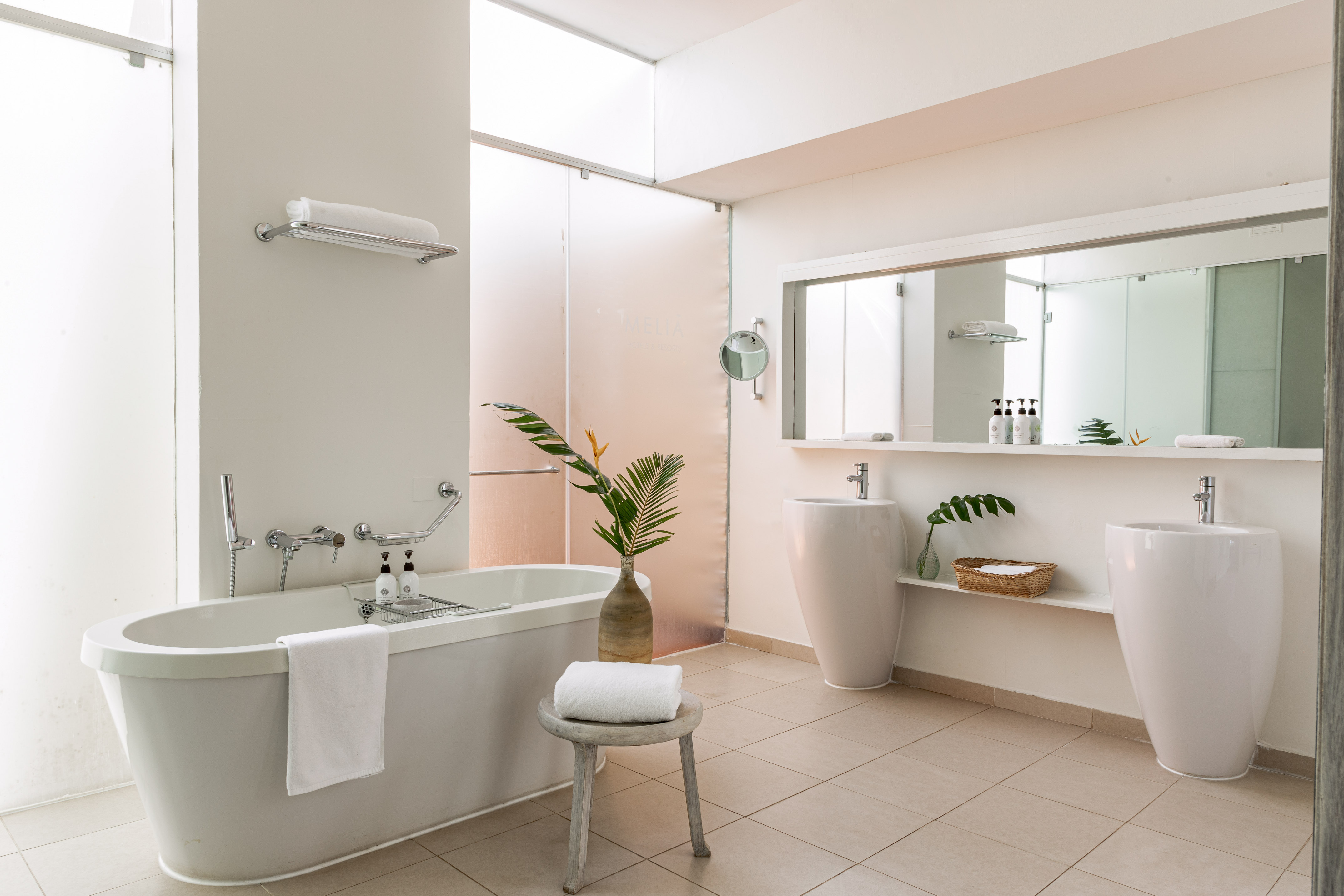 Modern hotel bathroom: freestanding tub, double sinks, tropical plant accent.