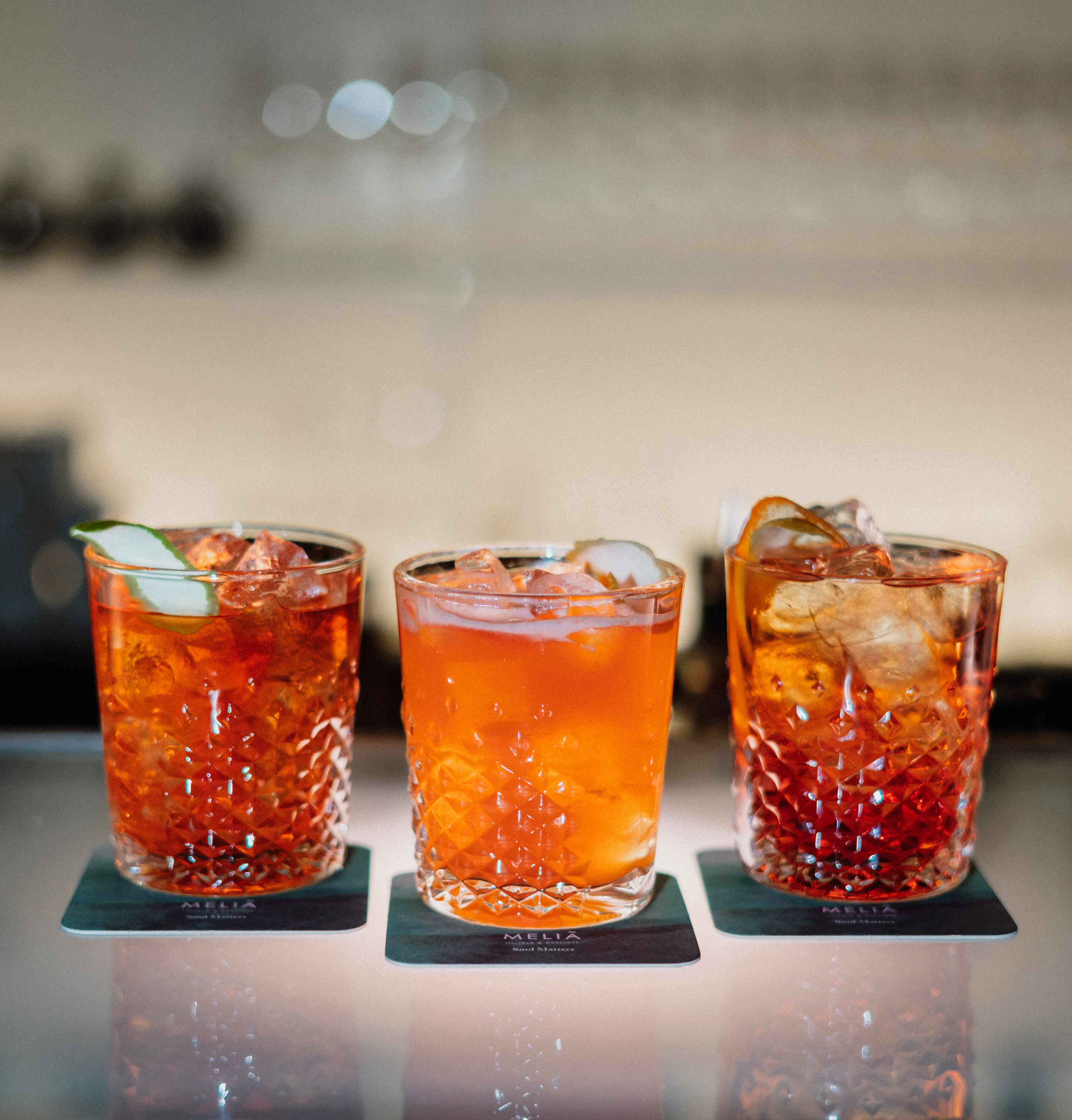Three orange cocktails with ice and garnishes on Meliã branded coasters.