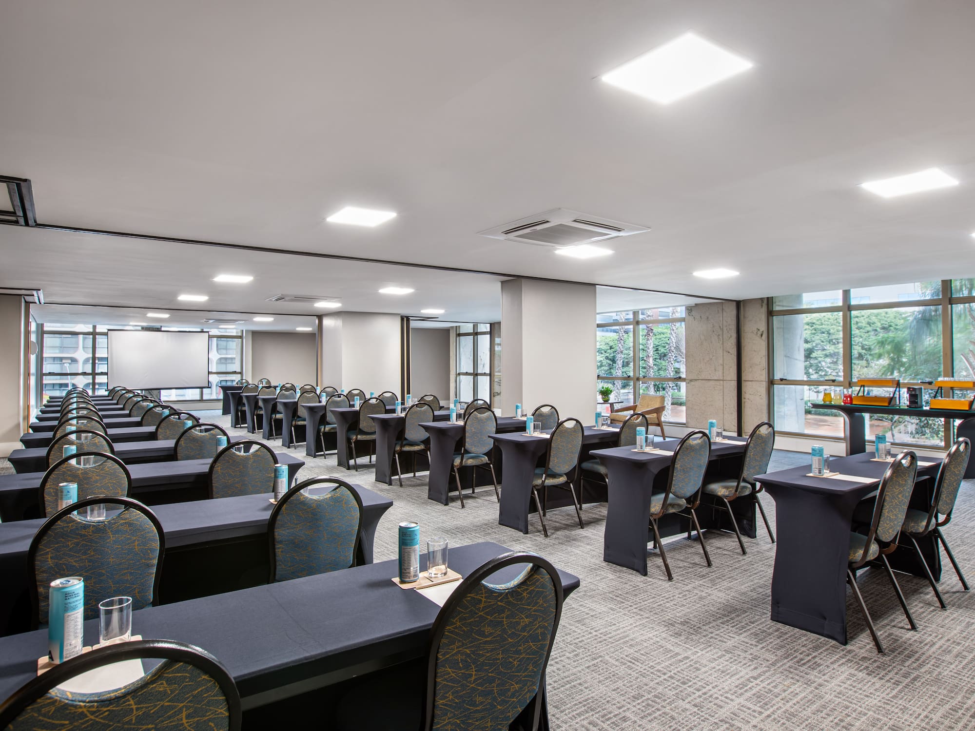 Empty conference room with tables, chairs, projector screen, and bright windows.