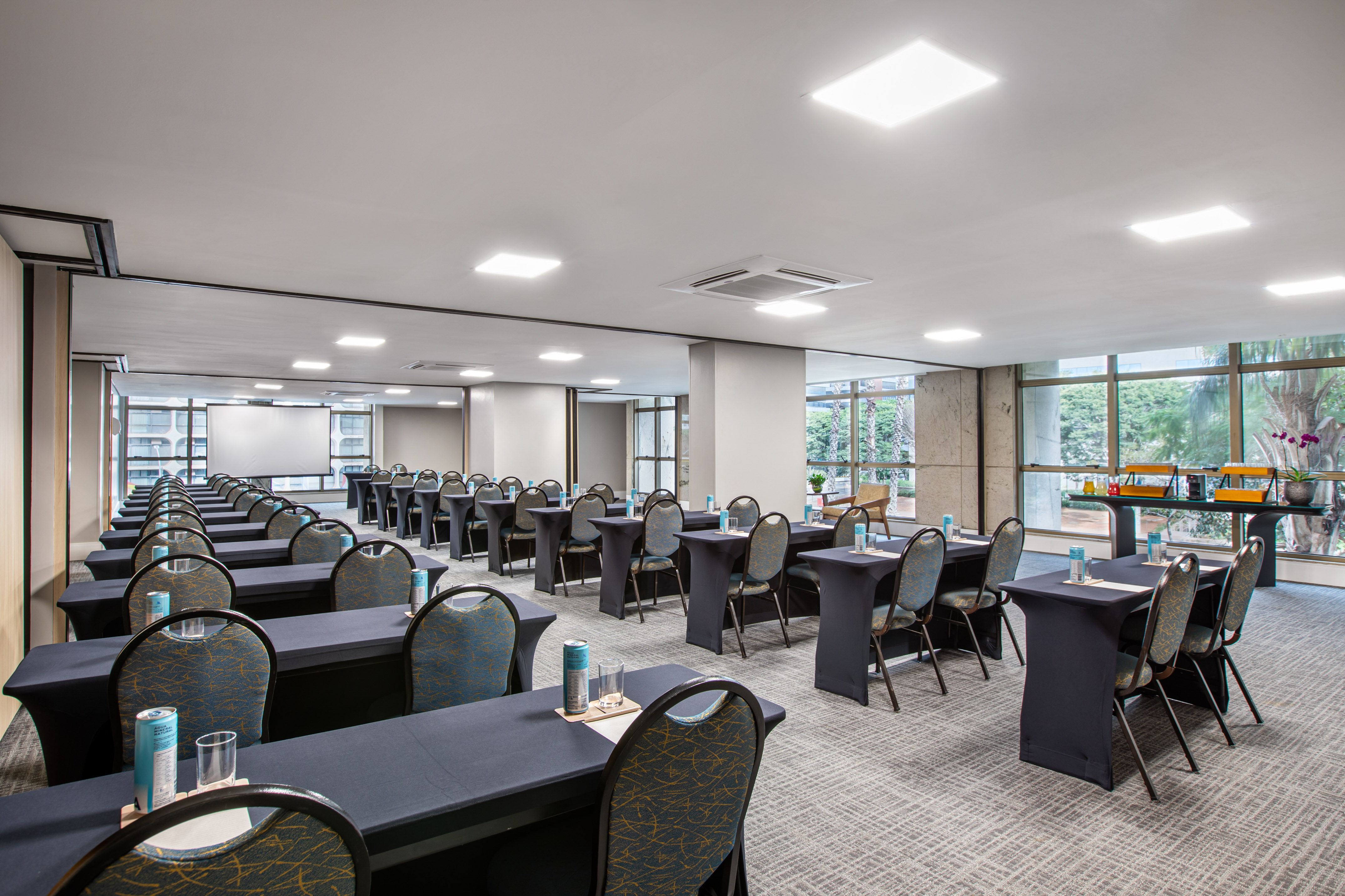 Hotel conference room setup with tables, chairs, projection screen, and natural light.