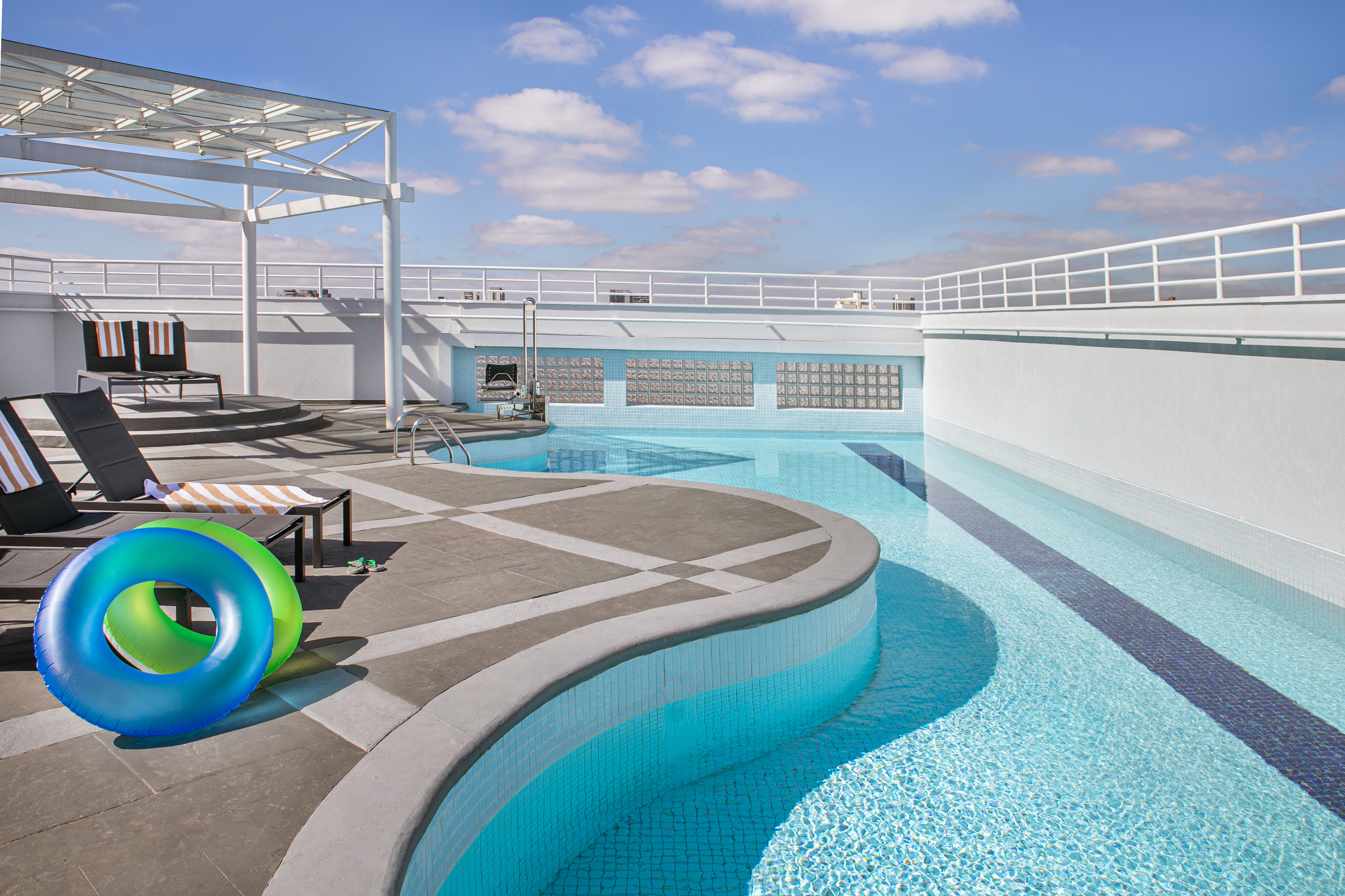 Rooftop swimming pool with lounge chairs, blue and green floats under a sunny sky.