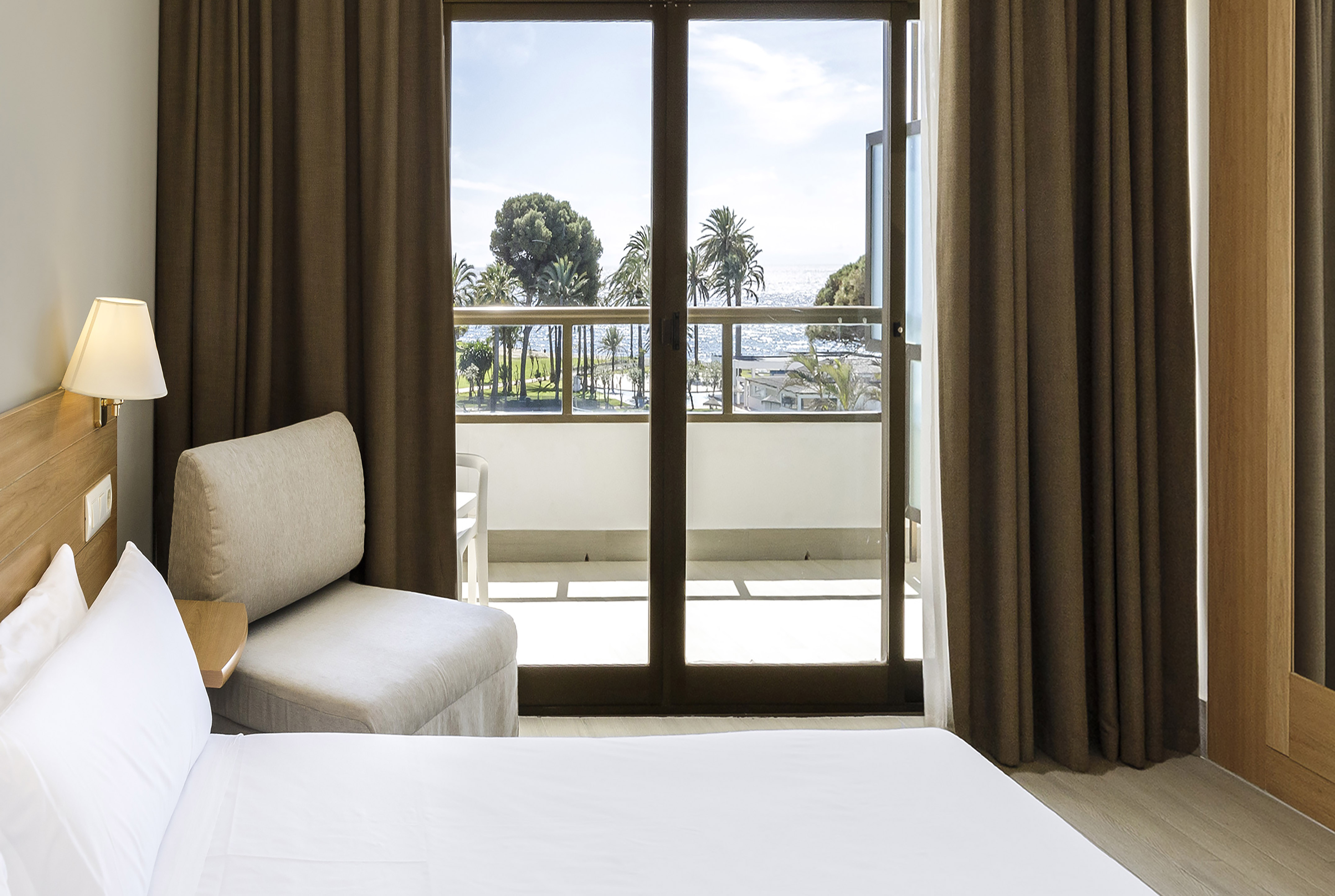 Hotel room with ocean view balcony, palm trees, bed, and armchair.