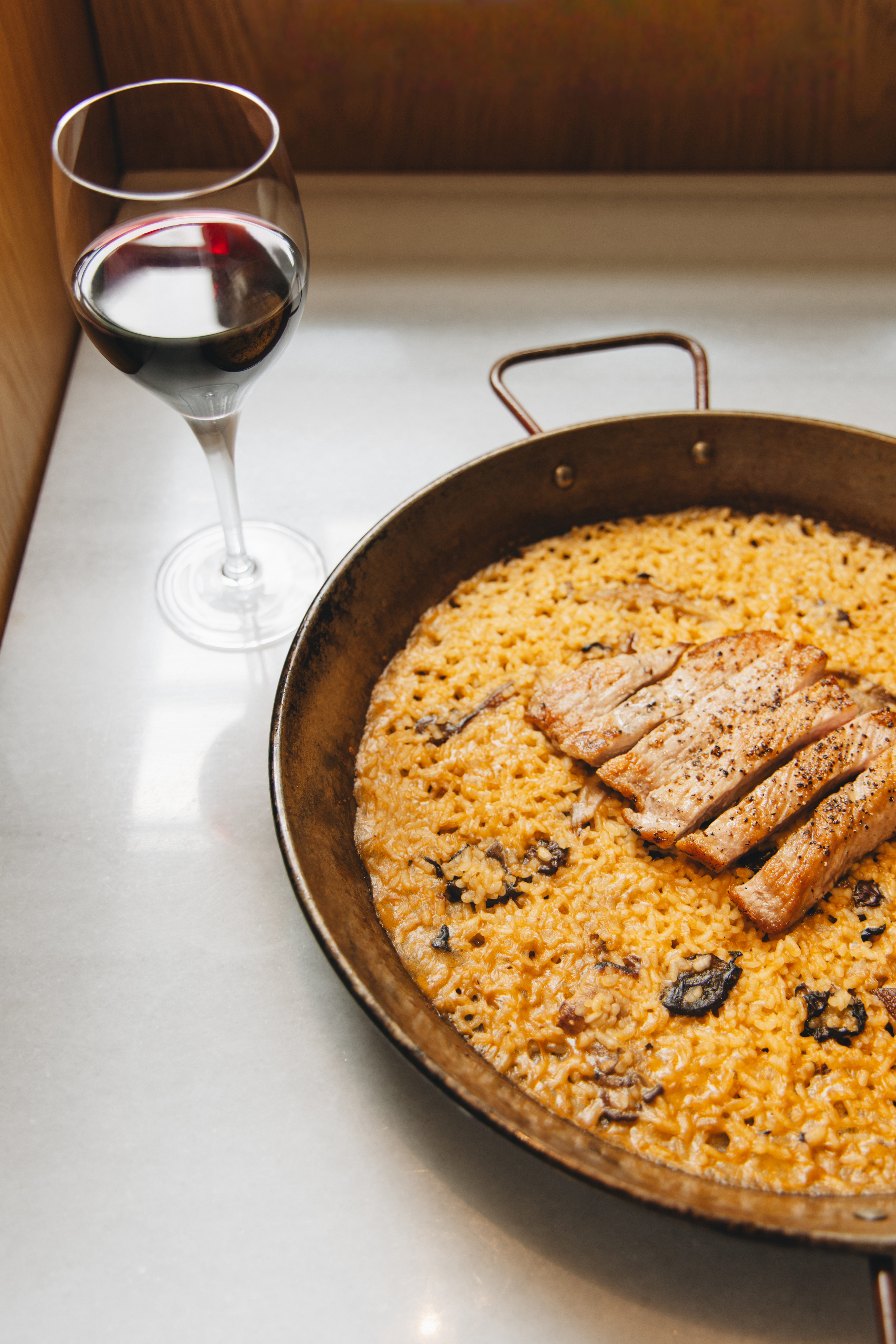 Golden pork paella with mushrooms, red wine glass on a white marble table.