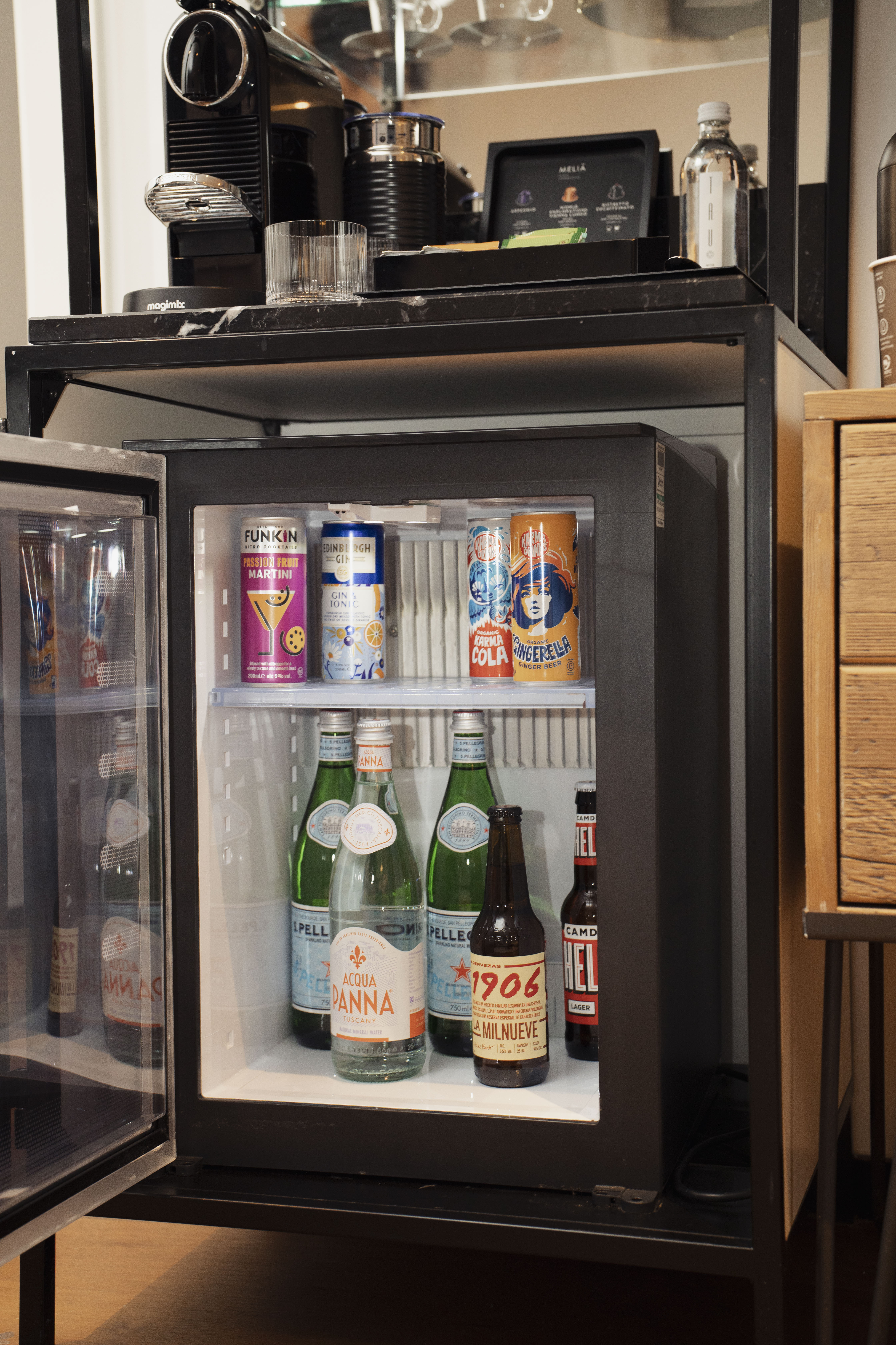 Hotel mini-bar with open fridge stocked with various drinks and Nespresso coffee machine.