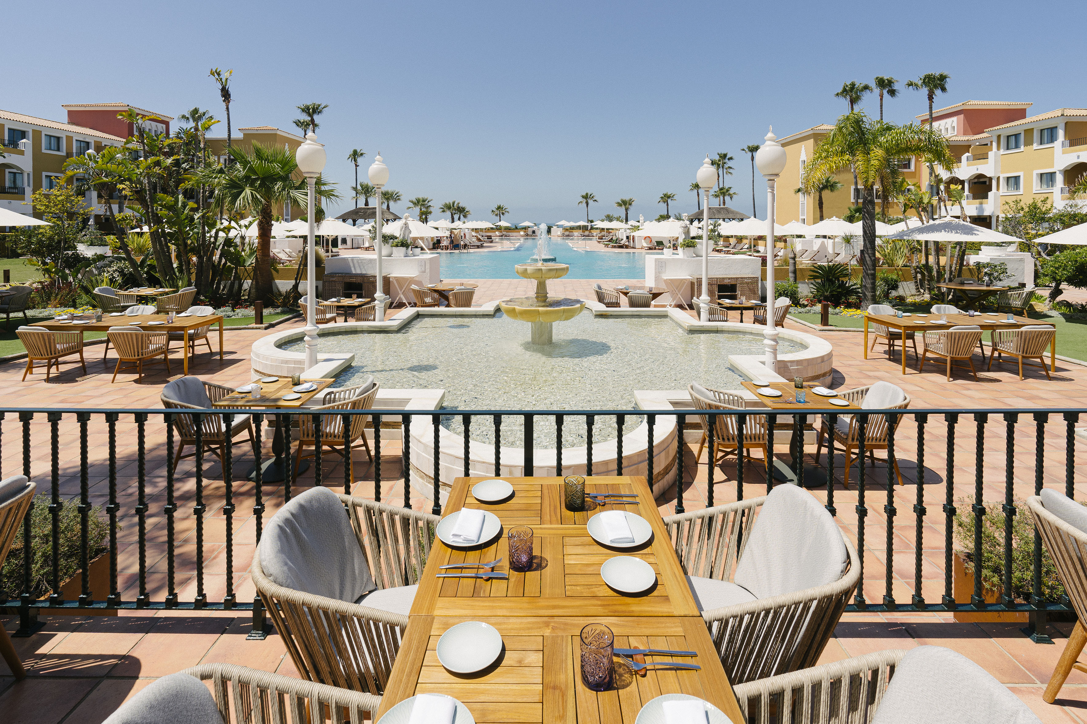 Sunny resort restaurant terrace with set tables, fountain, large swimming pool, and palm trees.