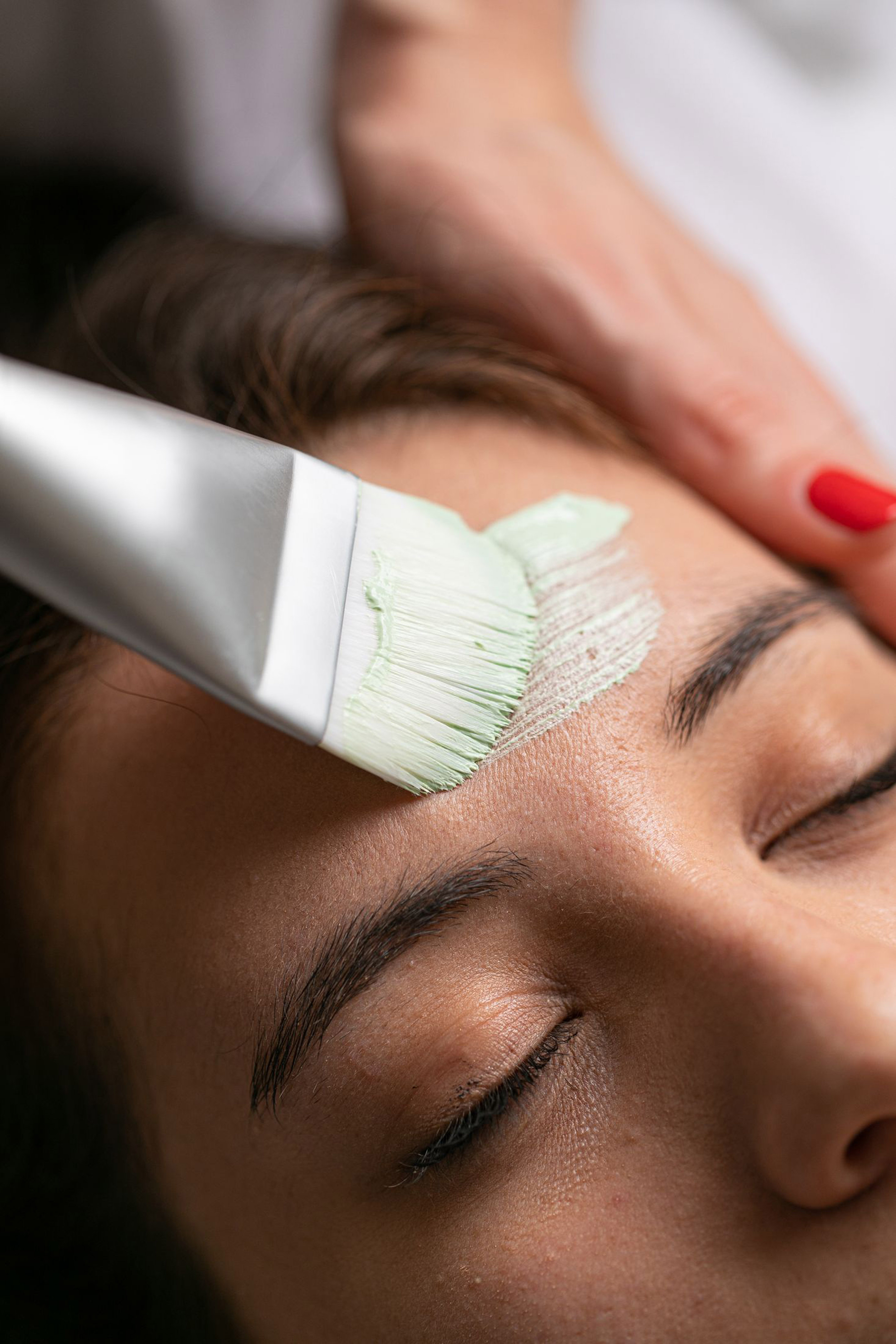 Brush applying green face mask to a person's forehead for skincare.
