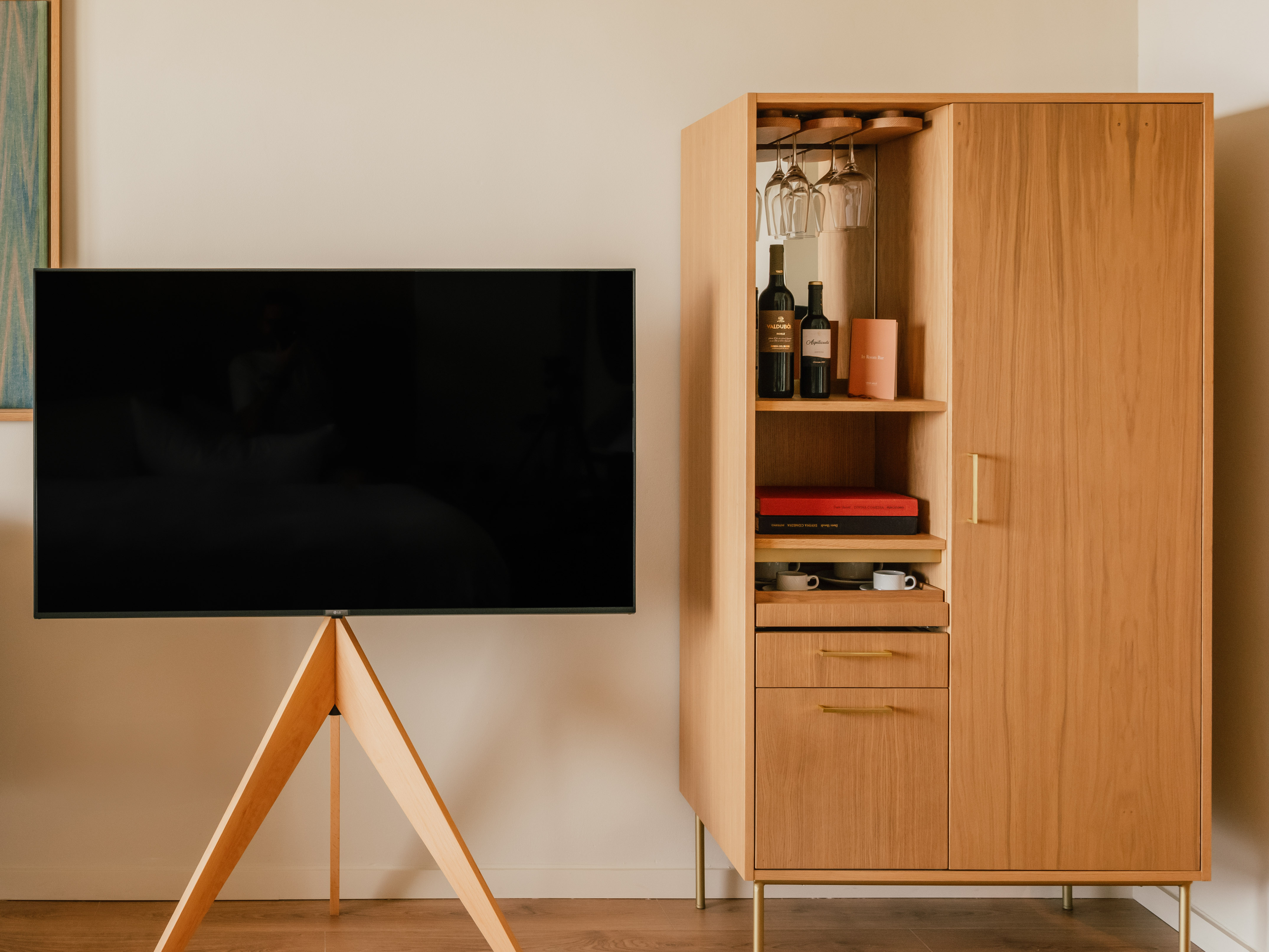 Flat-screen TV on wooden tripod beside a modern wooden minibar cabinet.